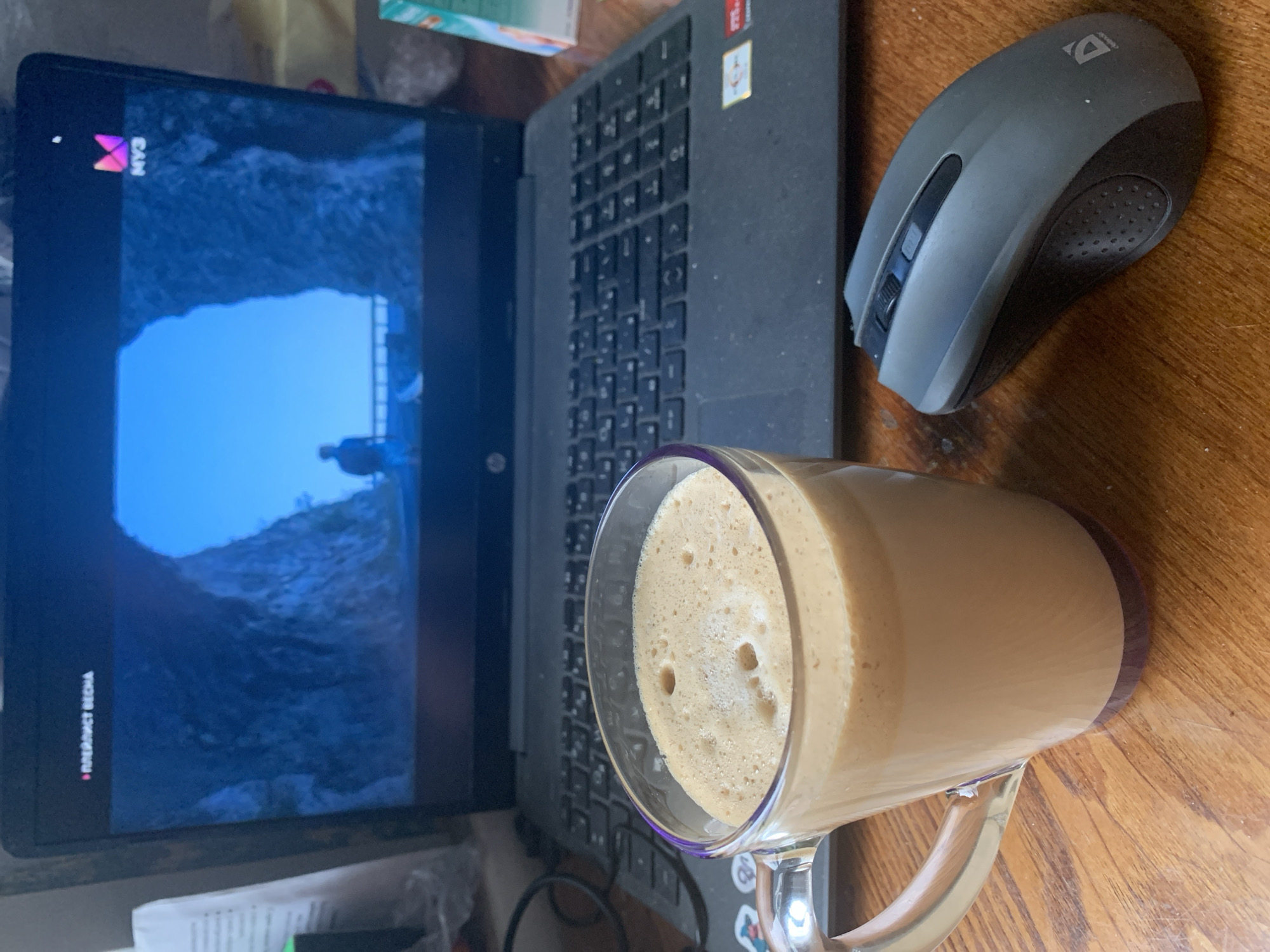 The image shows a laptop, a mug filled with a coffee beverage, and a computer mouse on a wooden surface. The laptop screen displays a scene of a tunnel with a person standing in the distance. The laptop's logo "AMD" is visible on the upper right corner. A "MY3" logo is also visible on the upper left of the laptop screen. The mouse is dark gray and labeled "Defender". The mug is clear and contains a frothy light brown beverage. Text that appears to be in Russian is visible on the bottom left of the screen.
Based on the text "ИЖЕВСКАЯ ВЕСНА", the laptop is showing content from Izhevsk.