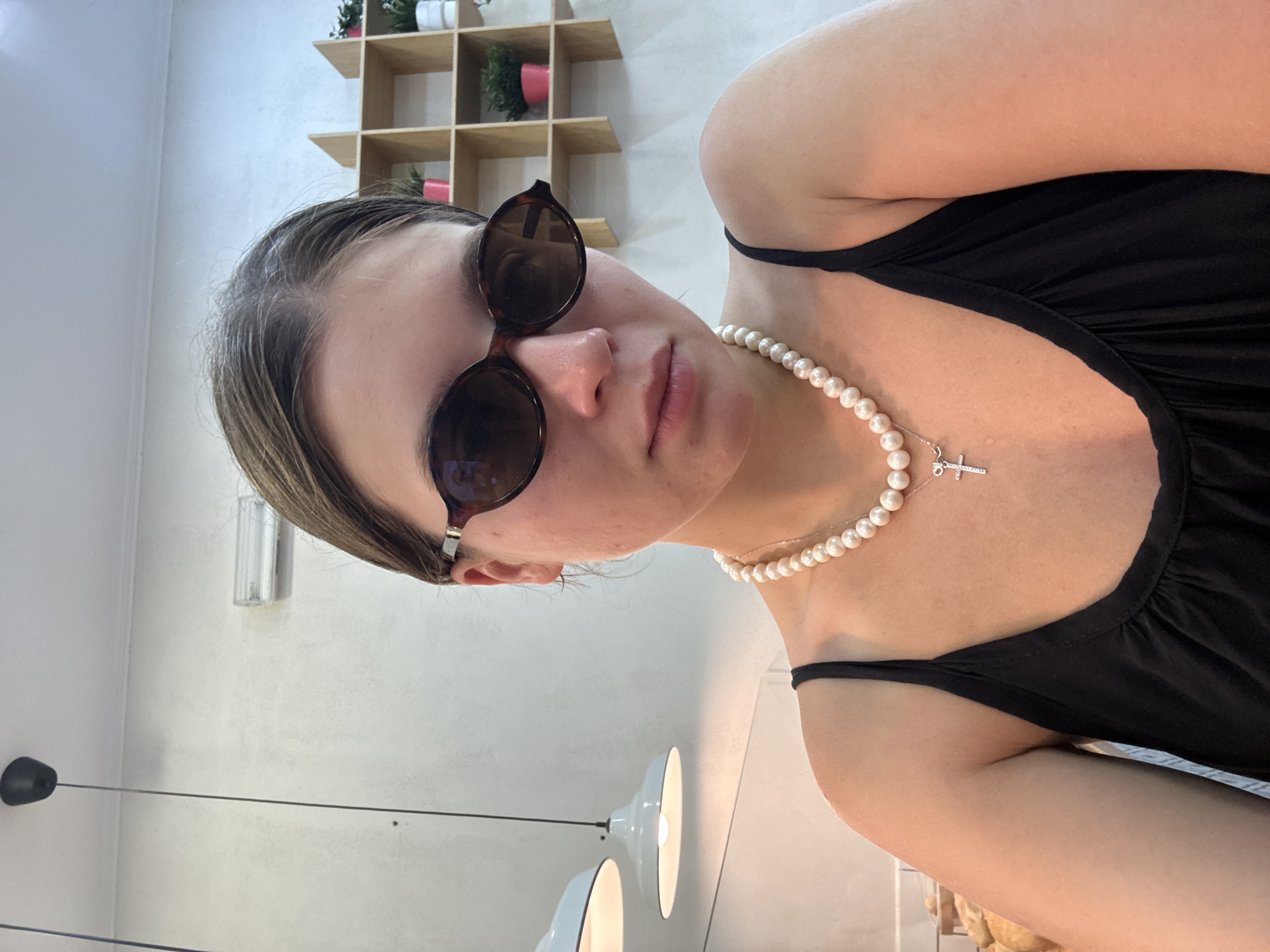 A medium close-up, eye-level shot shows a young woman indoors, wearing dark sunglasses, a black spaghetti strap top, and a pearl necklace with a silver cross pendant. Her hair is pulled back. She has light skin with a few blemishes. In the background, a light-colored wall features a wooden shelf unit with several potted plants. To the left, two pendant lights hang from the ceiling. The overall environment suggests a casual indoor setting, possibly a cafe or shop. There is no indication of the image being taken in Belgrade, Serbia.