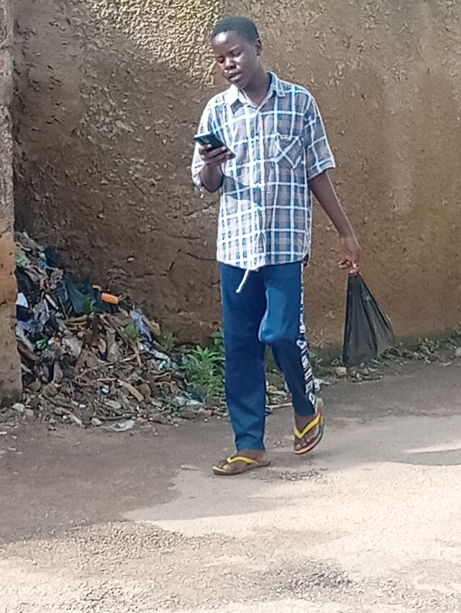 A young man wearing a blue and white checkered short-sleeved shirt, blue track pants with white lettering, and flip-flops is walking on a paved surface. He is holding a black plastic bag in his right hand and looking at his phone, which he holds in his left hand. To his left is a pile of refuse, including plastic bags and debris, against a rough, textured wall. Green vegetation is visible in the background. The scene appears to be an outdoor environment in a city like Jos, Nigeria, possibly a roadside or alleyway with accumulated waste.