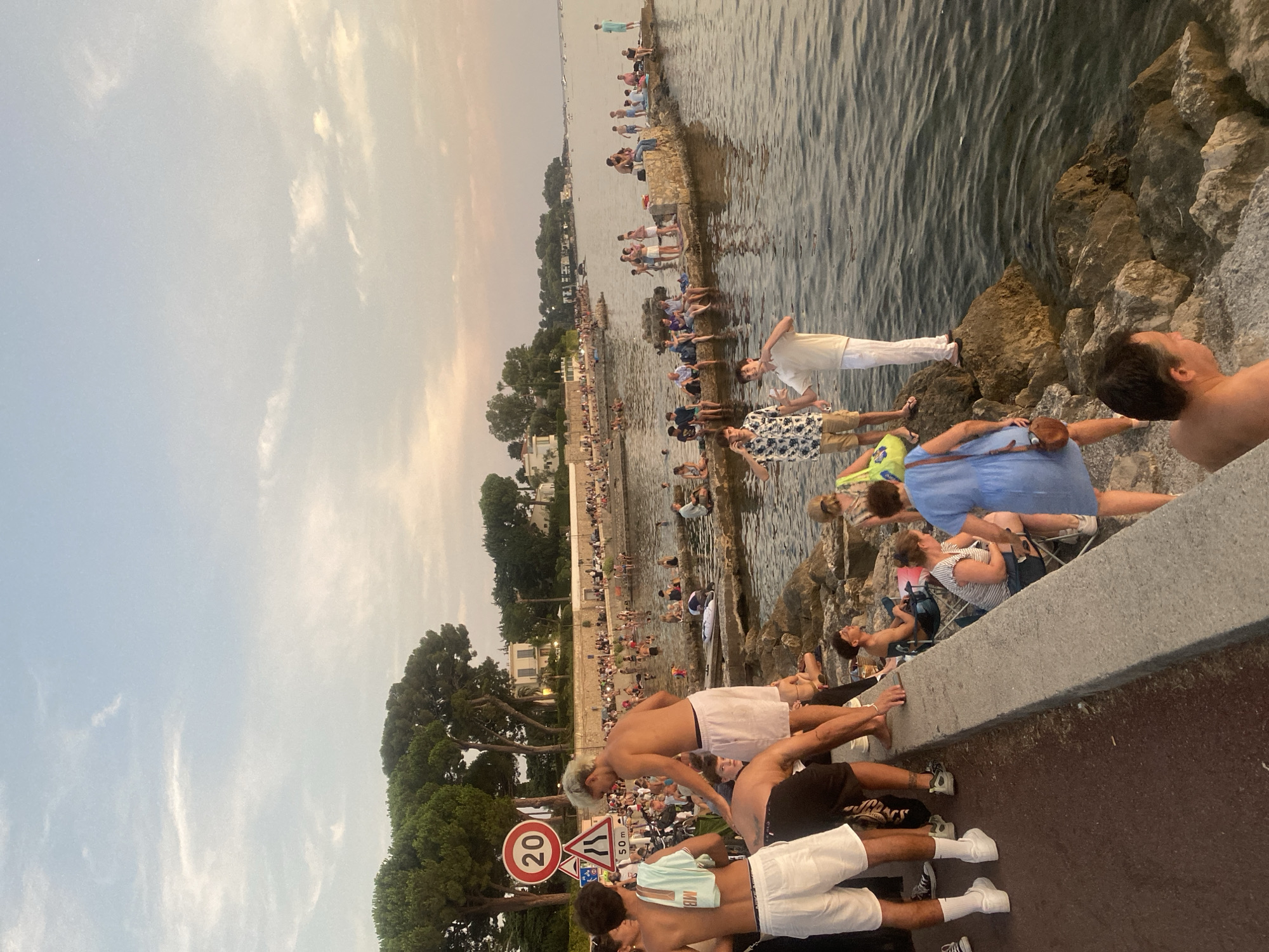 This is a vertical, eye-level, medium shot of a crowded waterfront area in Antibes, France, during the late afternoon or early evening. The scene depicts a seaside promenade with people gathered along a concrete barrier and on rock formations near the water. The sky is partly cloudy with a soft, warm light, suggesting either sunrise or sunset.
In the foreground, several young men are visible. On the left, one man is wearing white shorts and a light blue t-shirt draped over his shoulder, with white sneakers. To his right, another man in white shorts and a white t-shirt leans forward, with his back to the camera. Slightly behind them, a man with blonde hair and white shorts is standing, facing the water. A man in a white shirt and white pants stands on a rock formation on the right, looking out at the water.
Further back, a diverse group of people are seated and standing along the waterfront and on rocky outcrops. Many are dressed in casual summer attire, and some are in swimwear. The water is calm with gentle ripples. In the background, lush green trees line the shore, with residential buildings visible behind them. A road sign indicating a 20 km/h speed limit and a 50-meter distance is also visible on the left. The overall atmosphere is one of leisure and social gathering by the sea.