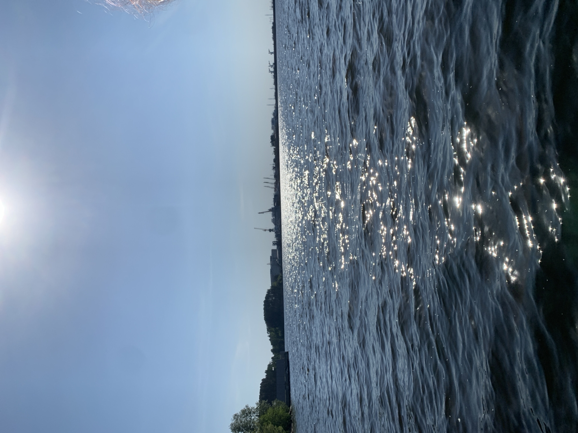 The image depicts a body of water, likely a river or lake, with sunlight reflecting off the surface, creating sparkling highlights. The water appears to have a slight ripple or wave pattern.
In the background, the skyline of a city is visible, including industrial structures like cranes. Along the water's edge, there are trees. The location is likely Chaikovsky, Russia, based on the presence of the industrial structures characteristic of a port city on the Kama River.
The sky is clear and blue, with the sun positioned to the left. A few hair strands or threads are visible near the top of the image.
