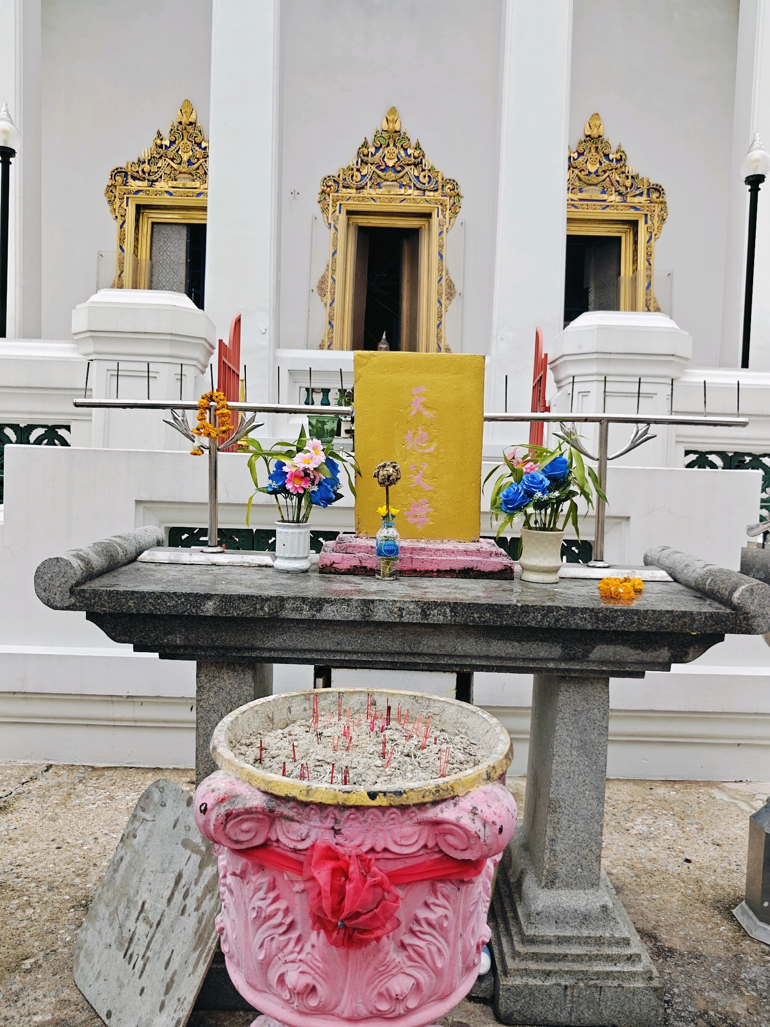 An outdoor altar is set up in front of a white temple building with ornate gold trim around three windows. The altar is made of granite and holds several items. In the center of the altar is a golden plaque with red Chinese characters, surrounded by pink and blue artificial flowers. A small glass bottle with blue liquid and a flower is placed in front of the plaque. To the right of the plaque is a small, orange flower garland.
In the foreground, a large, pink urn filled with ashes and red incense sticks is situated on the ground. A red ribbon is tied around the urn. To the left of the urn, a grey, rectangular stone slab leans against the urn. The ground is paved with concrete. The temple building in the background features white pillars and a white railing.