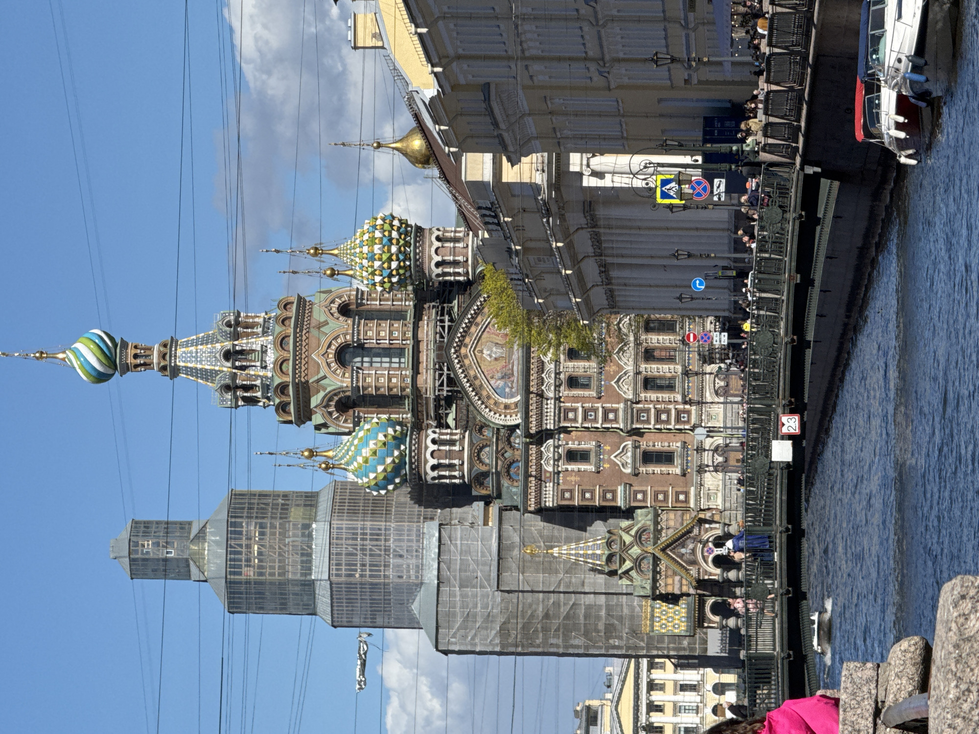 The image shows the Church of the Savior on Spilled Blood in Saint Petersburg, Russia, viewed from across a canal. The church is characterized by its colorful onion domes. Part of the building is covered in scaffolding and a protective structure. A waterway runs along the right side of the image. A group of people are visible along the canal's edge, as well as along the waterfront, where there are railings, buildings, lampposts, and signs. A boat with a Russian flag is also seen in the canal. Power lines are strung across the sky above the church.