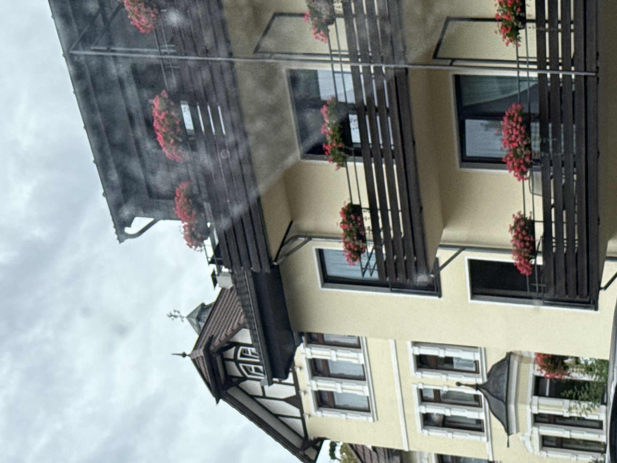 The image shows a multi-story building with a pale yellow facade, possibly a hotel or apartment building, located in the city of Bad Sachsa, Germany. The architecture features dark wooden balconies adorned with numerous planters filled with vibrant pink flowers. One section of the building exhibits a prominent gabled roof with dark timber framing and white infill, characteristic of some German architectural styles. The sky is overcast, suggesting a cloudy day. The foreground of the image is partially obscured by what appears to be water droplets or a streaky effect on the lens or window through which the photo was taken. No people are visible in the frame.