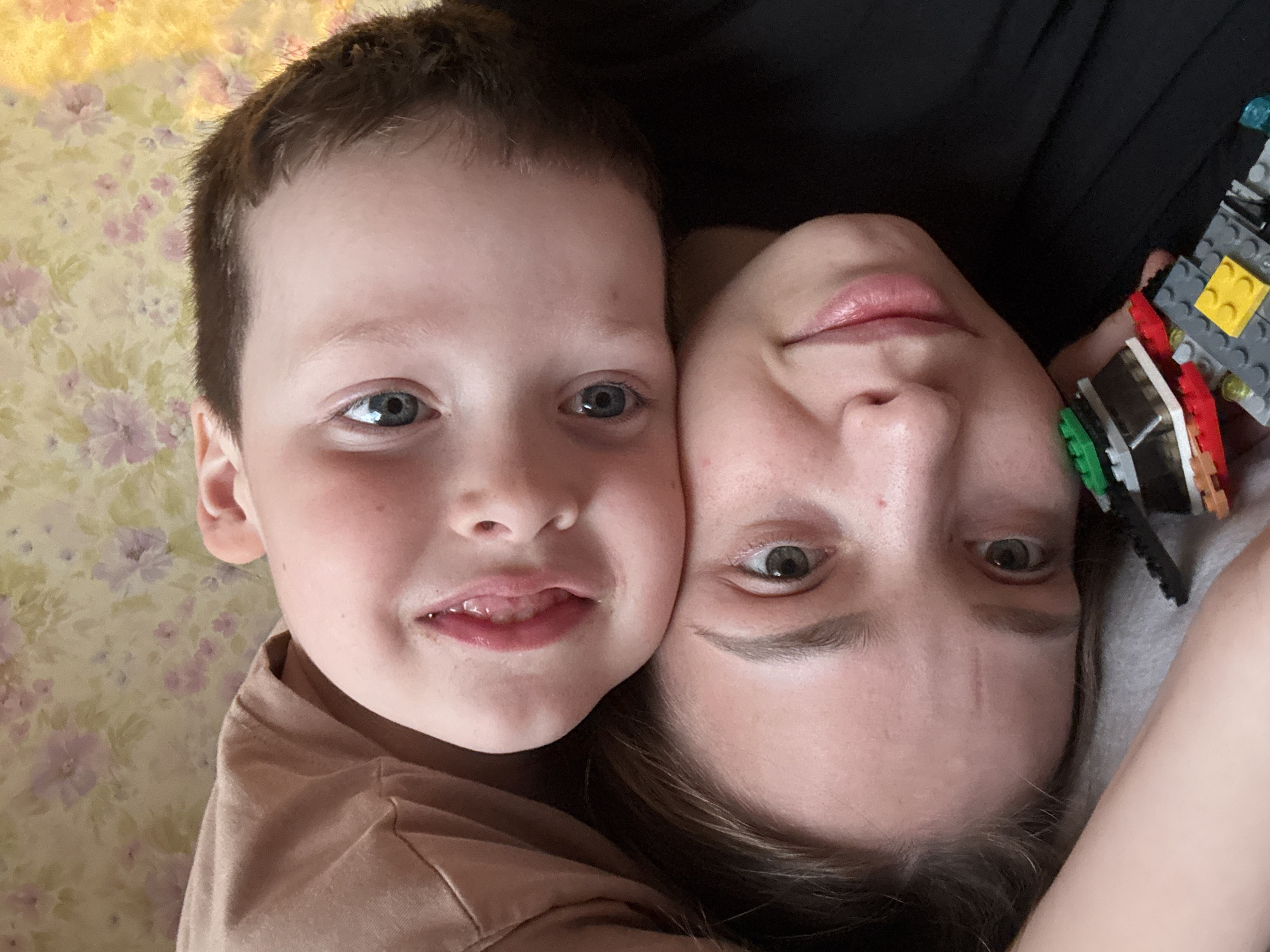 A close-up, eye-level shot shows a fair-skinned child and a fair-skinned adult posing closely together. The child is on the left, wearing a tan shirt, and has short brown hair and blue eyes. The adult, possibly female, is on the right, with brown hair and hazel eyes. The adult is holding a Lego construction. The background shows a floral patterned surface to the left of the child and a dark surface behind the adult. The faces of the child and the adult are close. There is no indication that this scene is in the city of Кемуль, Russia.