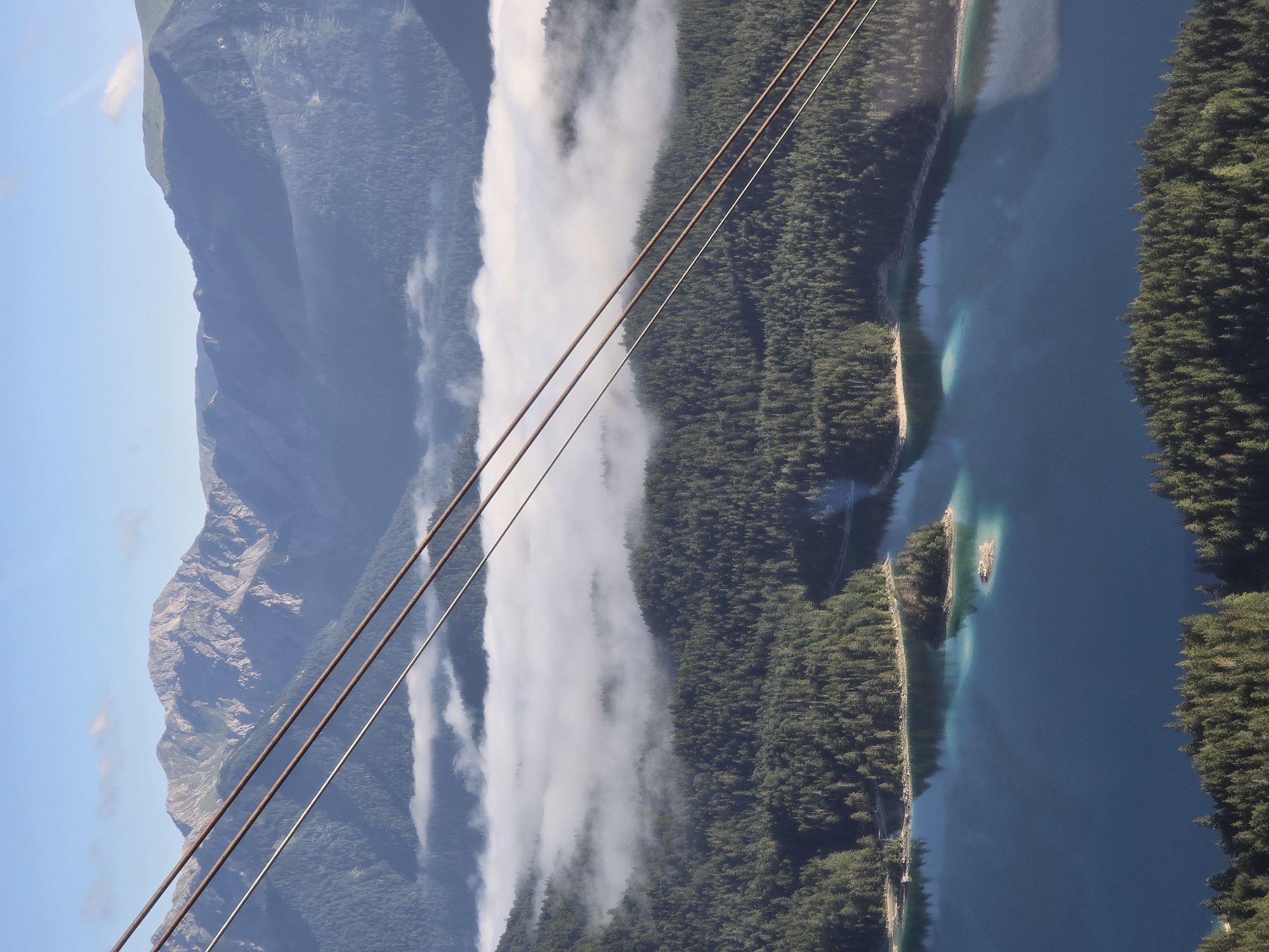 This image captures a breathtaking aerial view of the Eibsee lake in Grainau, Germany, with the Zugspitze mountain range dominating the background. Three parallel cable car lines traverse the foreground diagonally, stretching from the bottom left to the top right of the frame.
The Eibsee, a vibrant turquoise body of water, is dotted with small, tree-covered islands. The lake is embraced by a dense, dark green forest that carpets the surrounding hills. In the mid-ground, a thick layer of white clouds hugs the tree line, obscuring the lower slopes of the mountains and creating a dramatic contrast with the clear blue sky above.
The Zugspitze mountain range rises majestically in the distance, its rugged peaks tinged with gray and brown, indicating rocky terrain. The sky is a bright, clear blue with a few wispy white clouds scattered across it. The overall scene is one of serene natural beauty, with the man-made cable car lines providing a sense of perspective and human presence within the vast alpine landscape.