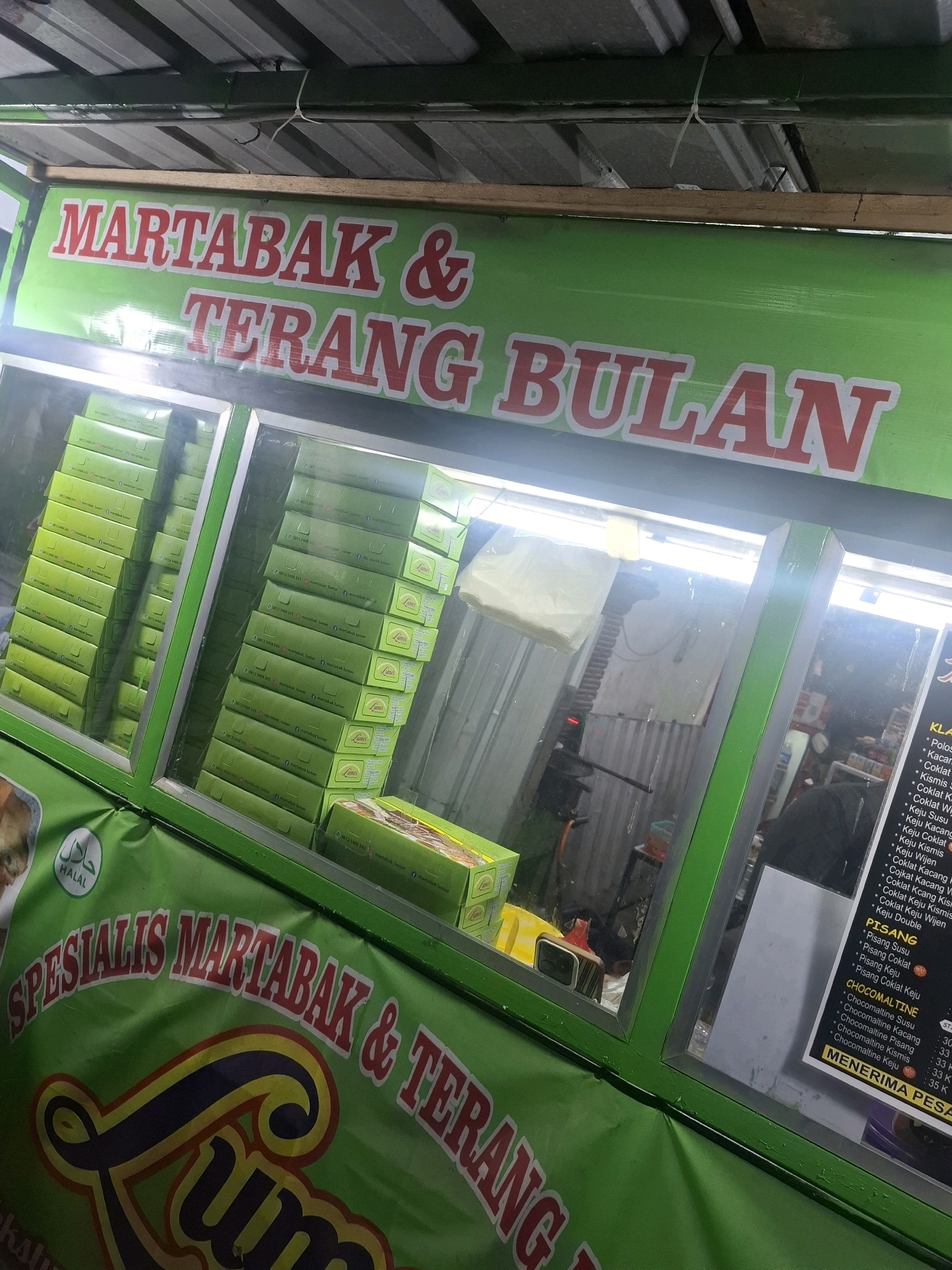 The image shows a brightly lit street food stall specializing in martabak and terang bulan, which are Indonesian sweet and savory pancakes. The stall is predominantly green with red lettering. The front of the stall prominently displays "MARTABAK & TERANG BULAN" in large red capital letters. Below this, a banner reads "SPESIALIS MARTABAK & TERANG" with a stylized "LUM" logo. The stall is stocked with numerous green boxes, presumably containing the finished products. A Halal certification symbol is visible on the left side. Through the glass display, a menu board is partially visible on the right, listing various toppings and flavors, such as "Keju Susu" (cheese milk) and "Coklat Kismis" (chocolate raisin). The background appears to be an outdoor, possibly alleyway setting, with corrugated metal walls and a visible ceiling structure. The lighting suggests it is either evening or an indoor/covered stall. The overall scene depicts a typical Indonesian street food vendor operation.