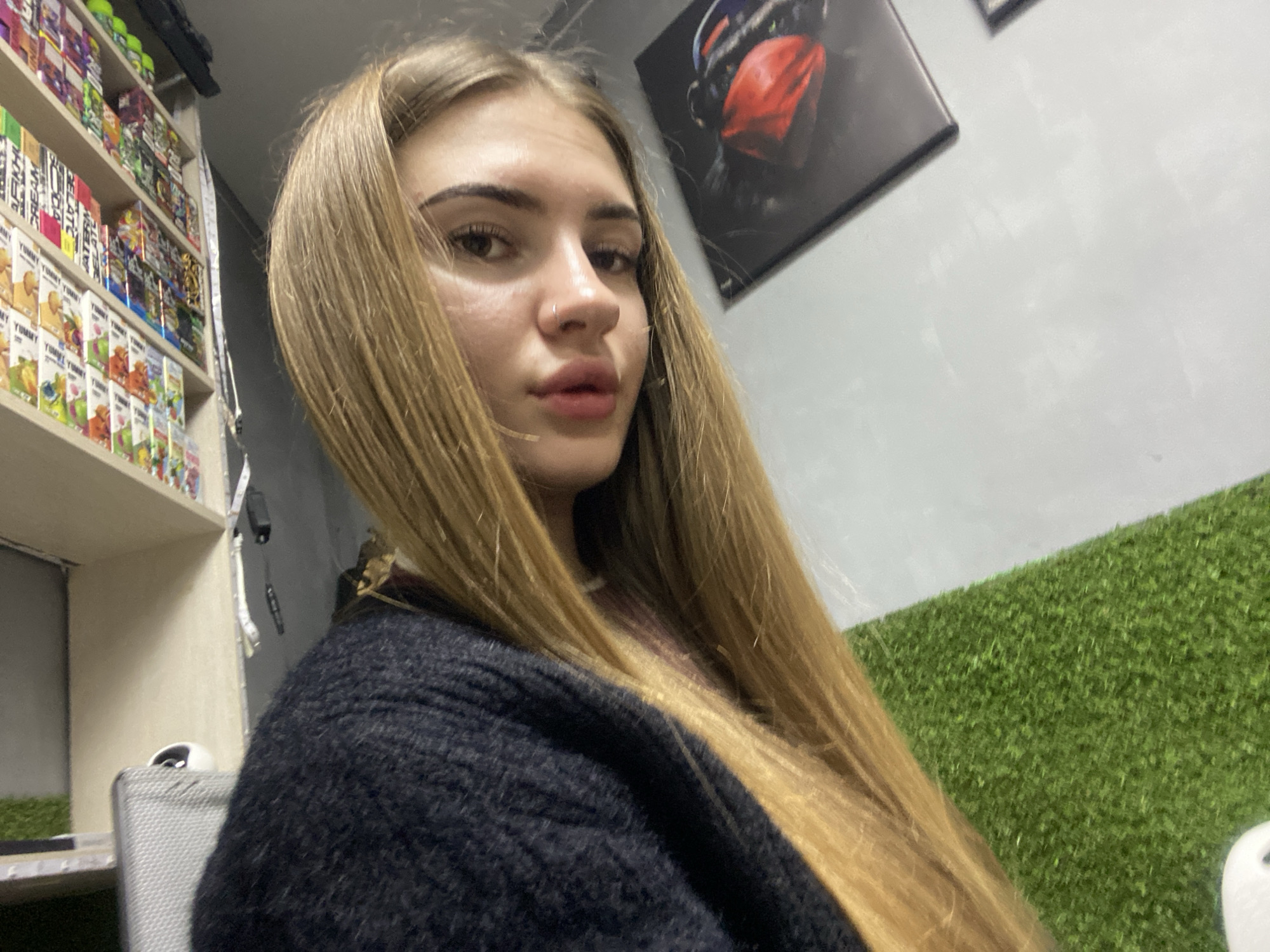 A selfie-style indoor shot captures a young woman with long, straight blonde hair looking directly at the camera. She has full, glossy lips and a small nose piercing on her left nostril. She is wearing a dark, possibly black, fuzzy knit cardigan or sweater. Her expression is neutral, with a slight pout.
The background reveals a retail or commercial setting. To the left, a tall, narrow shelf is packed with numerous small, brightly colored product packages. Text on some of the packages on the lower shelves clearly reads "YUMMY YUMMY," repeated multiple times, suggesting food or vape-related products. Other partially visible text includes "CREAM" and "WATERMELON." A measuring tape hangs loosely from the side of this shelf.
On the right, a light gray wall features a framed picture depicting a dark-furred monkey-like figure with a vibrant red, possibly fabric, mask or bandana covering its face. Below the picture, to the lower right, a section of artificial green grass (turf) is visible, extending towards the bottom of the frame. A small, white rounded object, possibly part of a chair or another fixture, is partially seen on the artificial grass. Part of a silver-colored chair is also visible behind the woman's left shoulder. The overall lighting suggests an indoor environment, possibly a shop or casual commercial space.