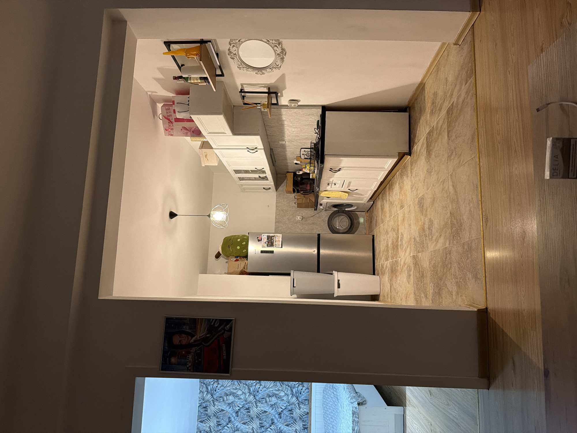 This is an indoor photograph of a residential kitchen and an adjacent bedroom, taken from a living room area. The kitchen features white cabinets, a black countertop, a silver refrigerator, and a washing machine. Above the cabinets, there are several decorative items and shopping bags. The floor is tiled in a neutral color. The bedroom, visible through an open doorway, has a white bed with patterned bedding and a wall with blue and white patterned wallpaper. The overall impression is of a functional, lived-in space. There is no indication of the city of Plovdiv, Bulgaria, or any specific forensic details in the image.
