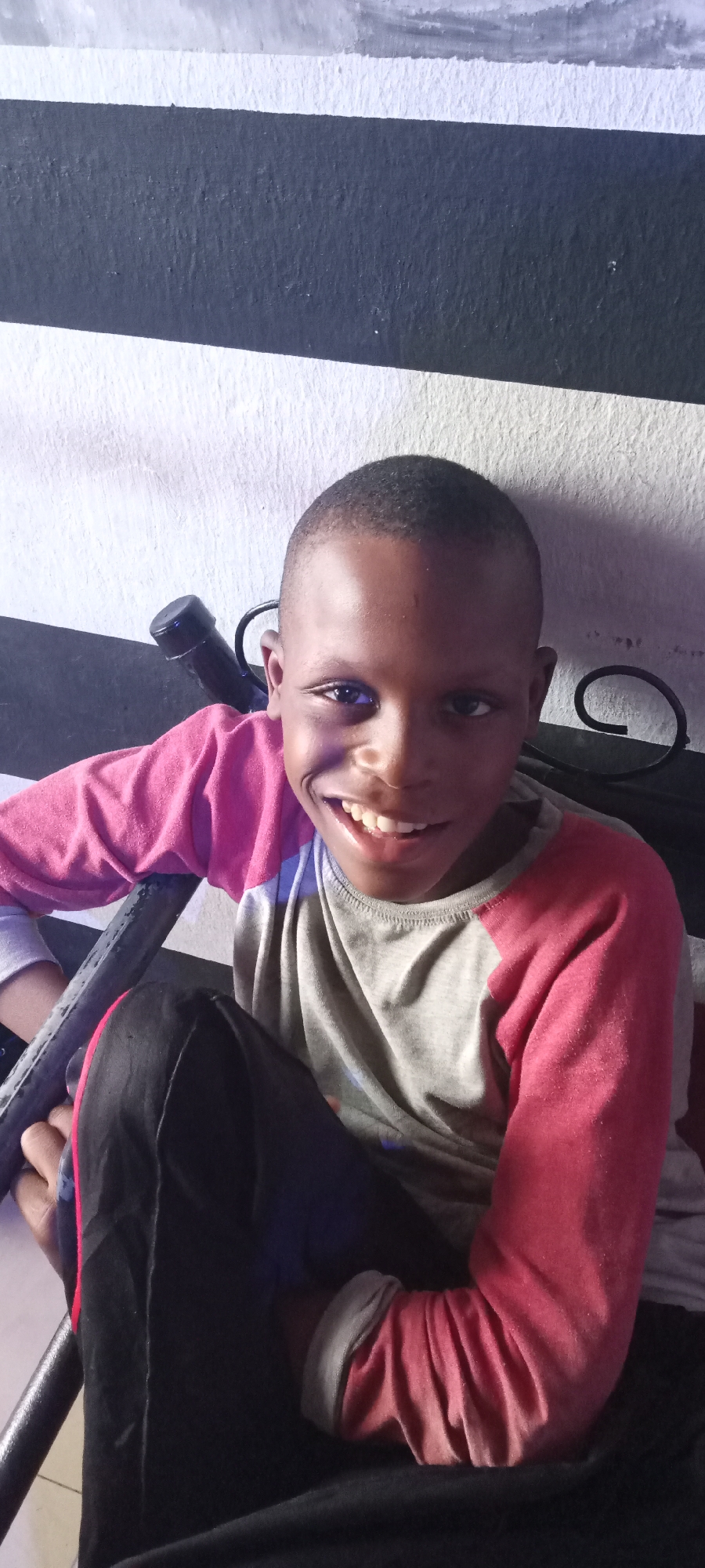 A young Black boy from Jos, Nigeria, is captured in a waist-up portrait, seated and smiling broadly directly at the camera. He is wearing a long-sleeved shirt featuring a grey torso and bright pink raglan sleeves, paired with black trousers. His short, dark hair is neatly cut, and his wide, joyful smile reveals his teeth, while his eyes appear bright and engaged. He is positioned slightly to the right of the frame, with his left arm resting on a black metal frame, possibly part of a chair, bed, or railing. The background is a wall with distinct horizontal black and white stripes, creating a bold, graphic pattern. The texture of the painted wall is visible, suggesting a textured plaster surface. The lighting appears to be indoor or shaded outdoor, highlighting the boy's face. The overall impression is one of youthful happiness and approachability. There is no visible text in the image.