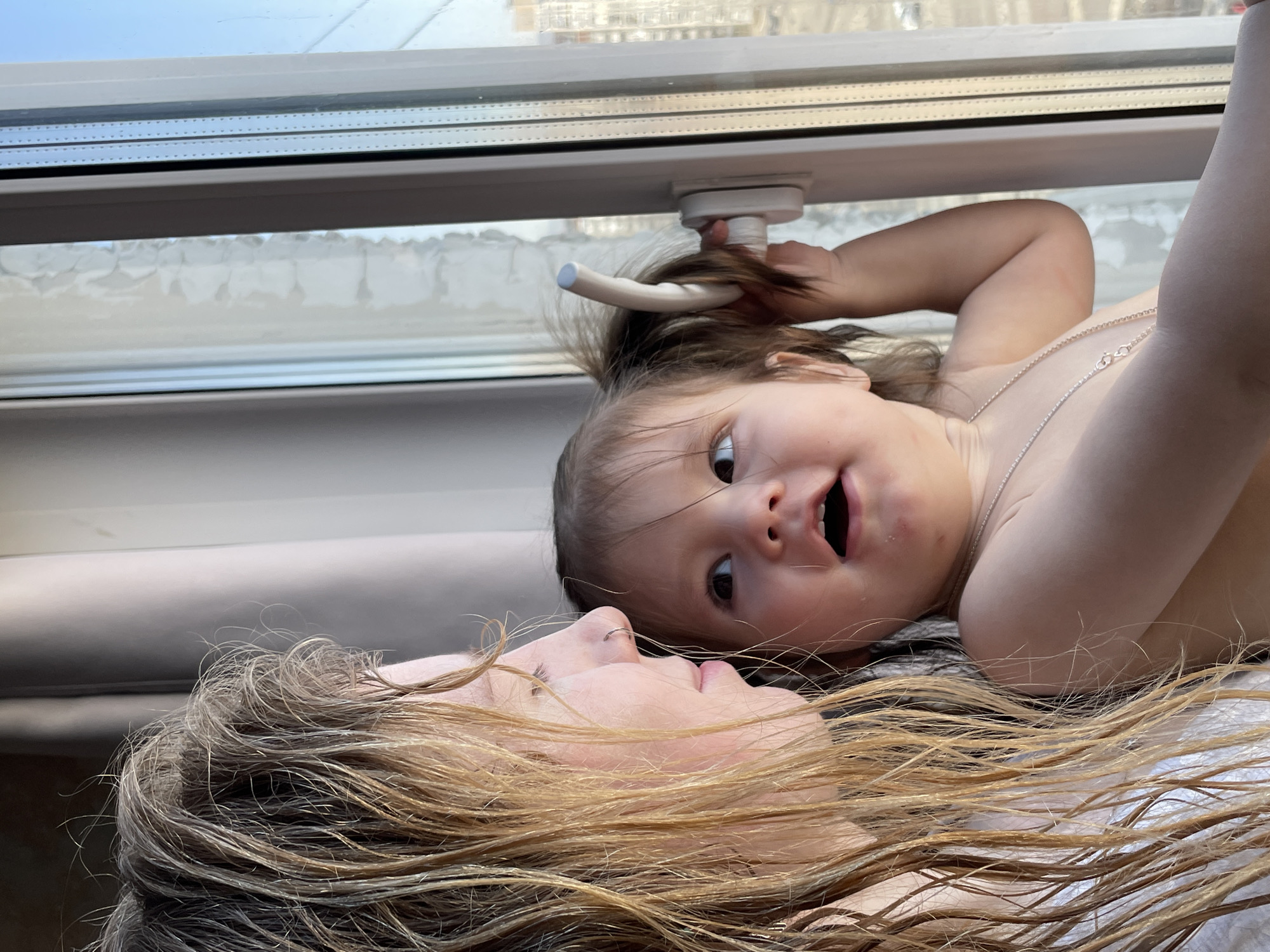 A mother and child lie on their backs near a window. The child, a baby, is closest to the window, holding onto what appears to be part of the window's mechanism. The mother is below the child. Both are looking at the camera. The woman has long blonde hair. The child is wearing a thin necklace. Through the window, an urban skyline is visible. The location is Izhevsk, Russia.