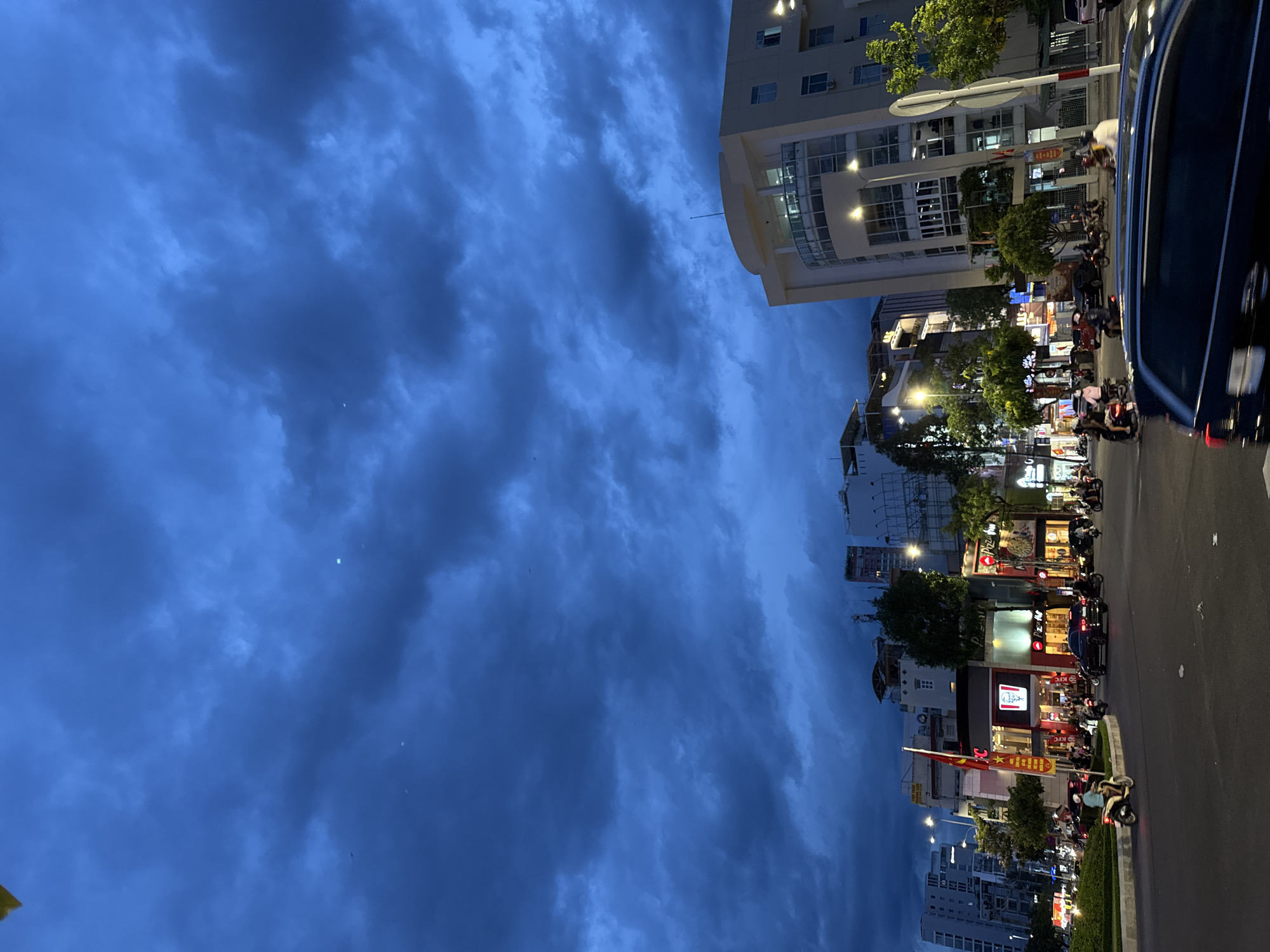 The image depicts a nighttime street scene in Nha Trang, Vietnam, under a dramatic, cloudy sky. The foreground shows a busy intersection with several vehicles, including motorcycles, cars, and a black SUV in motion. Pedestrians are visible on the sidewalks. The background features a commercial district with various buildings, including a KFC and a Pizza Hut restaurant illuminated by neon lights. The sky is a deep blue, with dark, textured clouds dominating the upper portion of the frame, suggesting an impending storm or twilight. Streetlights and building lights illuminate the scene, creating a mix of bright and shadowed areas. The overall impression is one of urban activity at dusk or night.