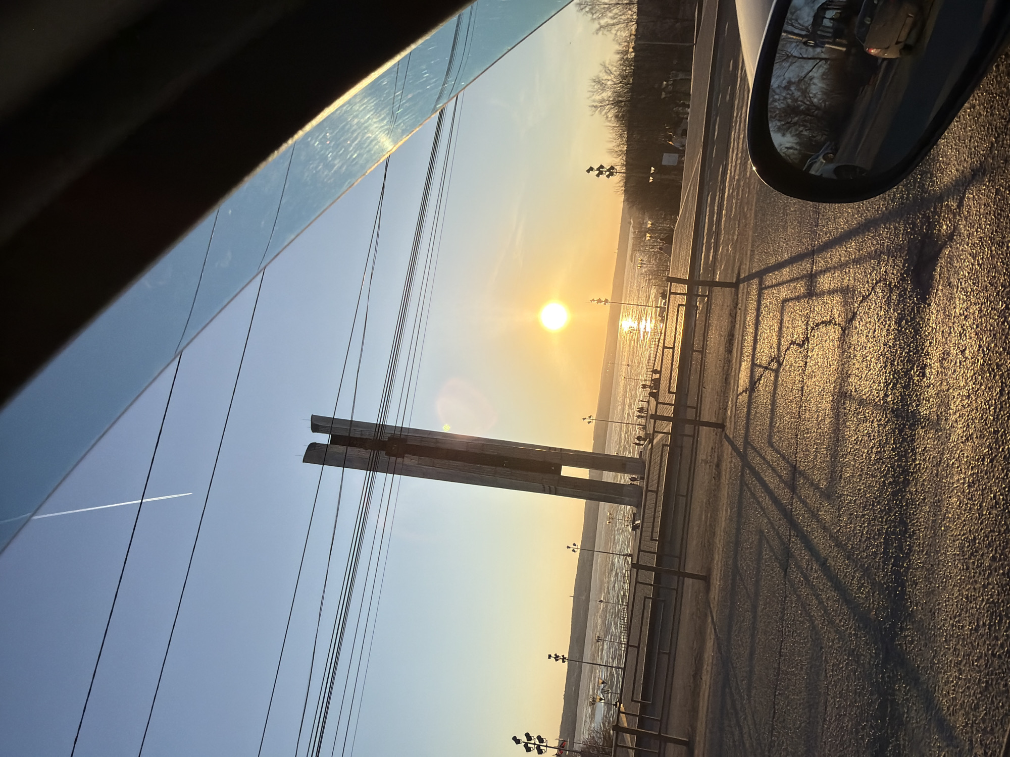 The image shows a view from inside a car in Izhevsk, Russia, overlooking a waterfront area at sunset. The setting sun reflects on the water, creating a golden glow. A prominent structure, possibly a monument or bridge support, stands near the waterfront. Power lines are visible against the sky. A contrail from an airplane is seen in the upper left. A side view mirror of the car with a vehicle reflected is visible on the right. The ground is paved, and shadows of the fence and streetlights are cast onto the ground. The view suggests a location along the embankment of the Izhevsk Pond.