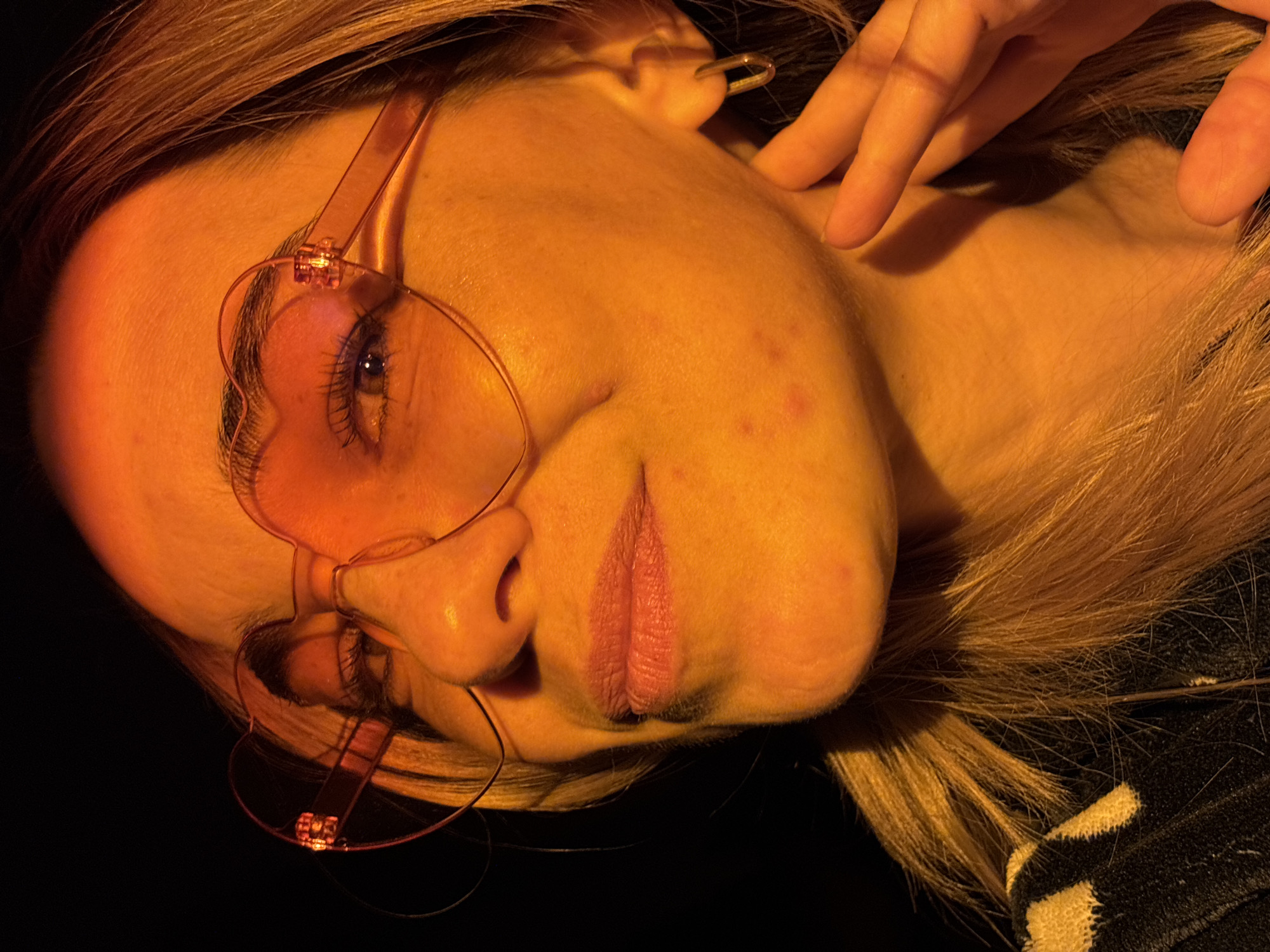 The image depicts a woman with blonde hair wearing heart-shaped glasses. The lighting is orange, possibly from artificial light. Her hand is near her neck. The background is dark. There is no indication of the location being in Unknown, Russia.