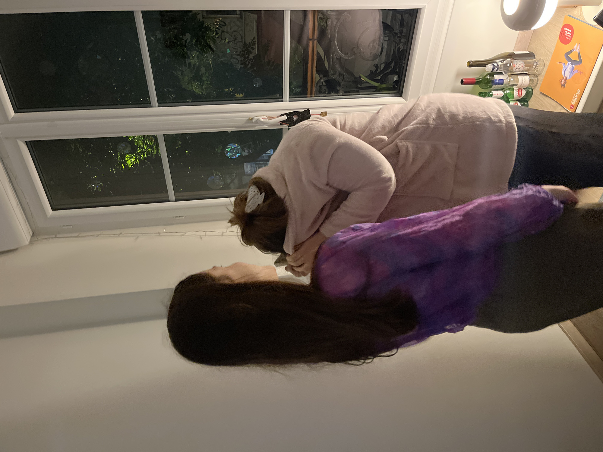 Two individuals are indoors near a window. One person is wearing a pink robe, the other is wearing a purple top. The window provides a view of foliage outdoors. Adjacent to the window, there are bottles and a book. The location is not identified.