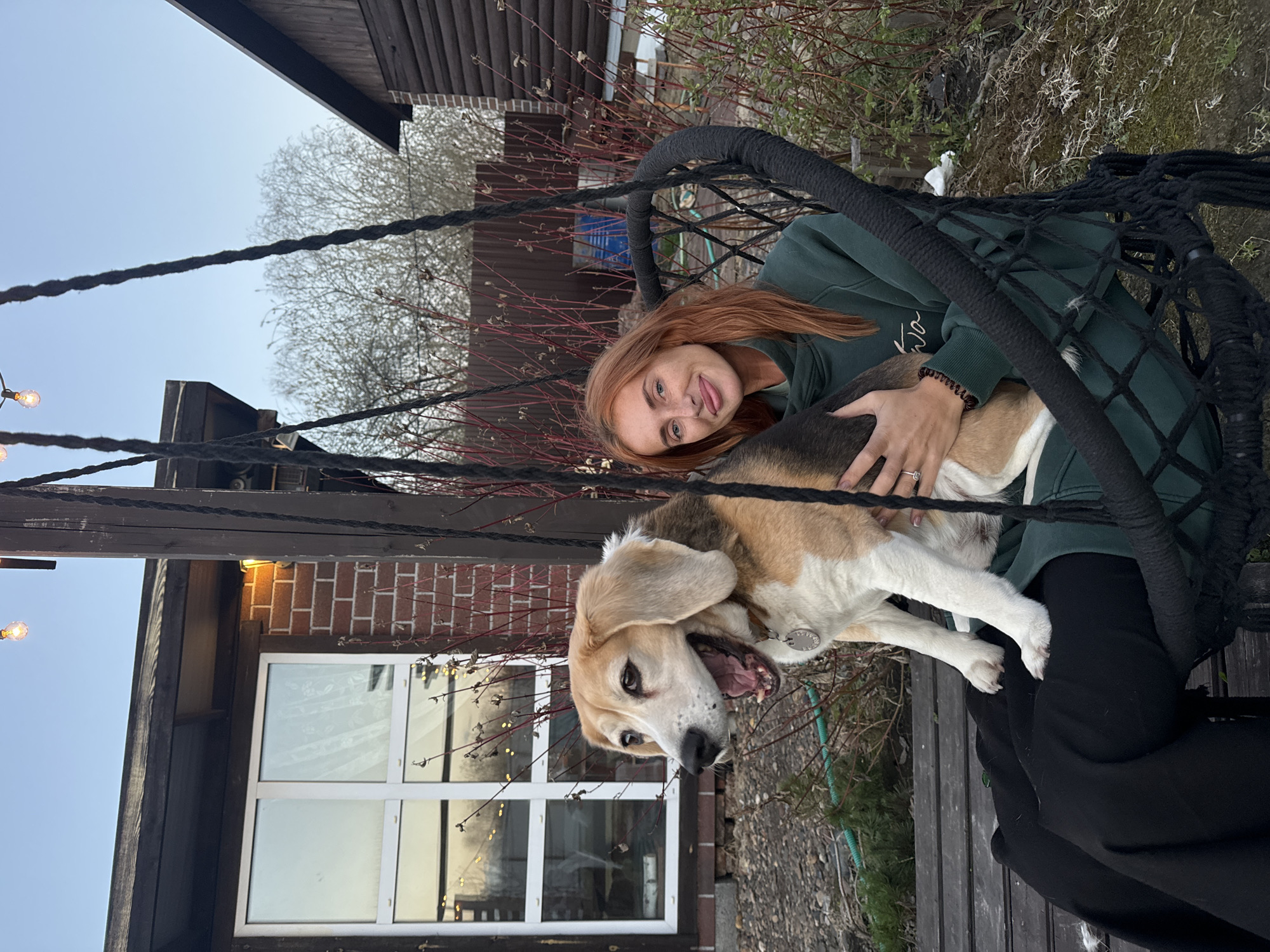 A woman and a beagle dog are in a black hammock swing. The woman has red hair and is wearing a green sweatshirt and black pants. She is holding the dog. The beagle is tan, white, and black. The background shows a building with a window and bare trees. The photograph appears to have been taken in Emel'yanovo, Russia.