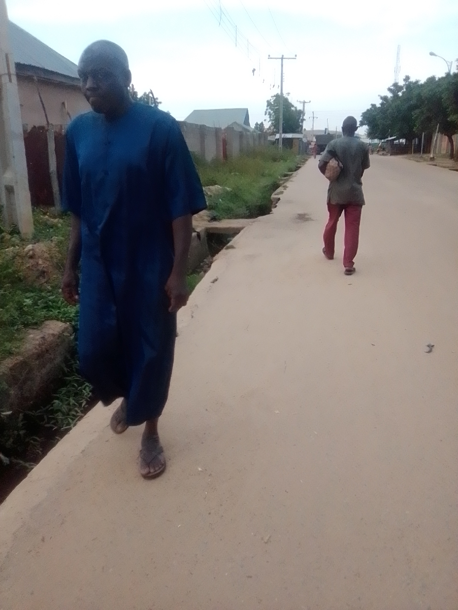 The image captures a street scene in Liji Hill, Nigeria. Two men are walking on a dusty, paved road. The man in the foreground, closer to the camera, is wearing a dark blue, loose-fitting garment and appears to be walking towards the viewer. He is barefoot. The second man is further down the road, walking away from the camera. He is wearing a green shirt and reddish-pink trousers and is carrying a light-brown, textured object under his left arm. The environment shows buildings with corrugated metal roofs and fences along the side of the road, with some vegetation visible. Power lines and poles are also present in the background. The sky is overcast.