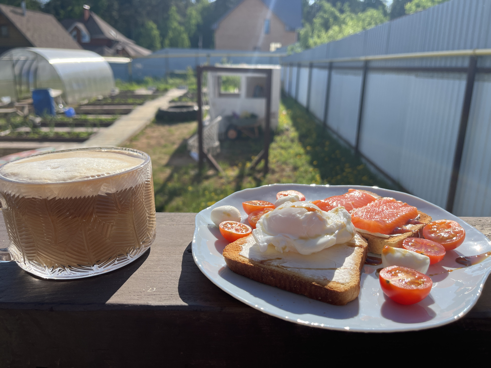 The image depicts a cup of coffee and a plate of food on a wooden surface, likely a balcony or porch. The food includes toast with cream cheese and a fried egg, salmon, cherry tomatoes, and small mozzarella balls, drizzled with a dark sauce.
In the background, there is a view of a residential area, with a greenhouse, a small shed, a fence, and other houses visible. The scene suggests a suburban or rural setting, bathed in daylight.
While the image is likely from a location in Russia, pinpointing it to Chaikovsky is not possible without additional information.