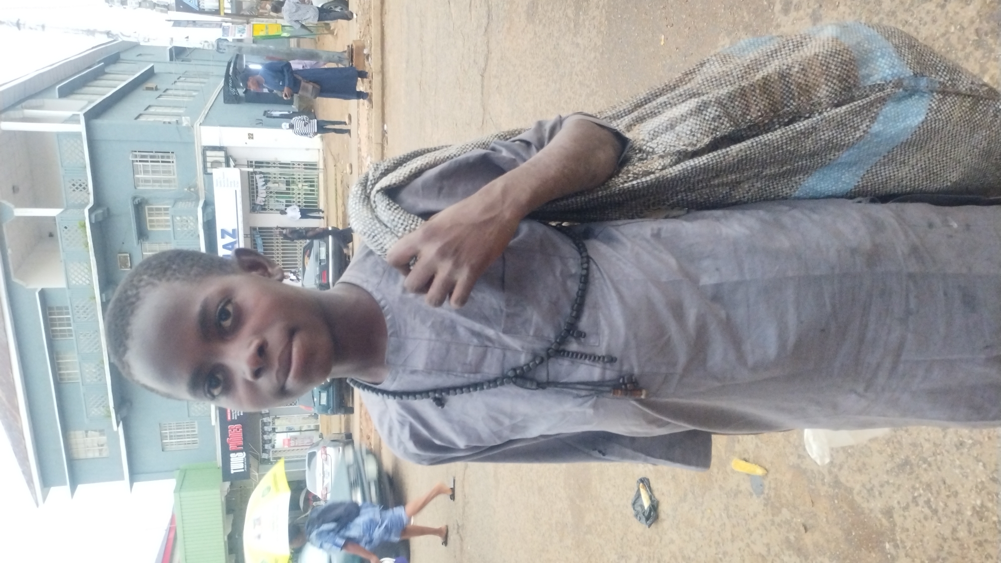 A young boy, appearing to be of African descent and approximately 10-12 years old, stands on a dirt and asphalt surface in what is identified as the city of Jos, Nigeria. He is wearing a plain, grey, long-sleeved traditional tunic. Around his neck, he wears a long strand of dark, wooden beads. He is carrying a large, woven sack over his right shoulder, which appears to be filled with goods. The background shows a multi-story building with a pale blue facade and balconies. Several other individuals are visible in the background, some walking and others standing near shops. Cars are present on the road behind the boy, suggesting a street or roadside setting. The lighting suggests it is daytime.