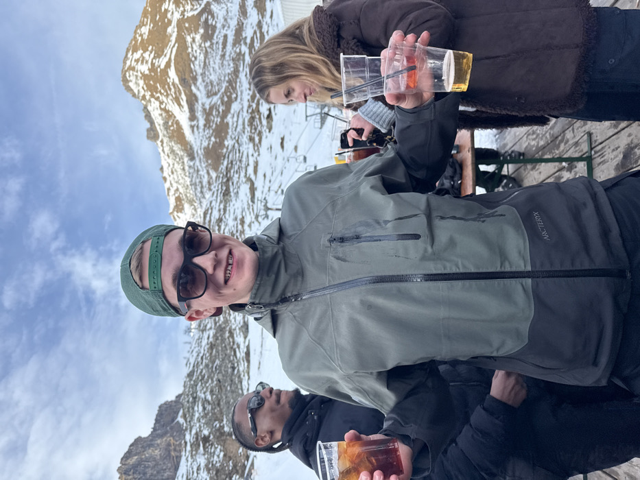 The image captures three individuals enjoying drinks outdoors on a bright, snowy day in a mountainous region, likely Alagna Valsesia, Italy.
In the foreground, a young man with light hair, wearing a green baseball cap backward and dark sunglasses, smiles broadly at the camera. He is dressed in an olive green and dark grey technical jacket, bearing the "ARC'TERYX" logo on the left chest area. He holds two clear plastic cups; one contains an iced, reddish-orange drink with a straw, and the other holds an amber-colored beverage.
Behind him, a woman with long, light brown hair is visible, wearing a brown, possibly fur-lined, winter jacket. She appears to be looking down or away from the camera, and her hand holds a smartphone. Partially visible in the lower right of the frame is a third person, a man in dark sunglasses and a black jacket, holding a clear plastic cup with a dark, iced drink.
The background reveals rugged, snow-dusted mountains under a clear blue sky with sparse white clouds. Structures of a ski lift are discernible between the subjects and the distant peaks, indicating a ski resort environment. The individuals are positioned on what appears to be a wooden deck or terrace. The overall mood is relaxed and social, suggesting an après-ski gathering or a break during a day on the slopes, with the bright sunlight and winter attire pointing to a crisp, cold day.