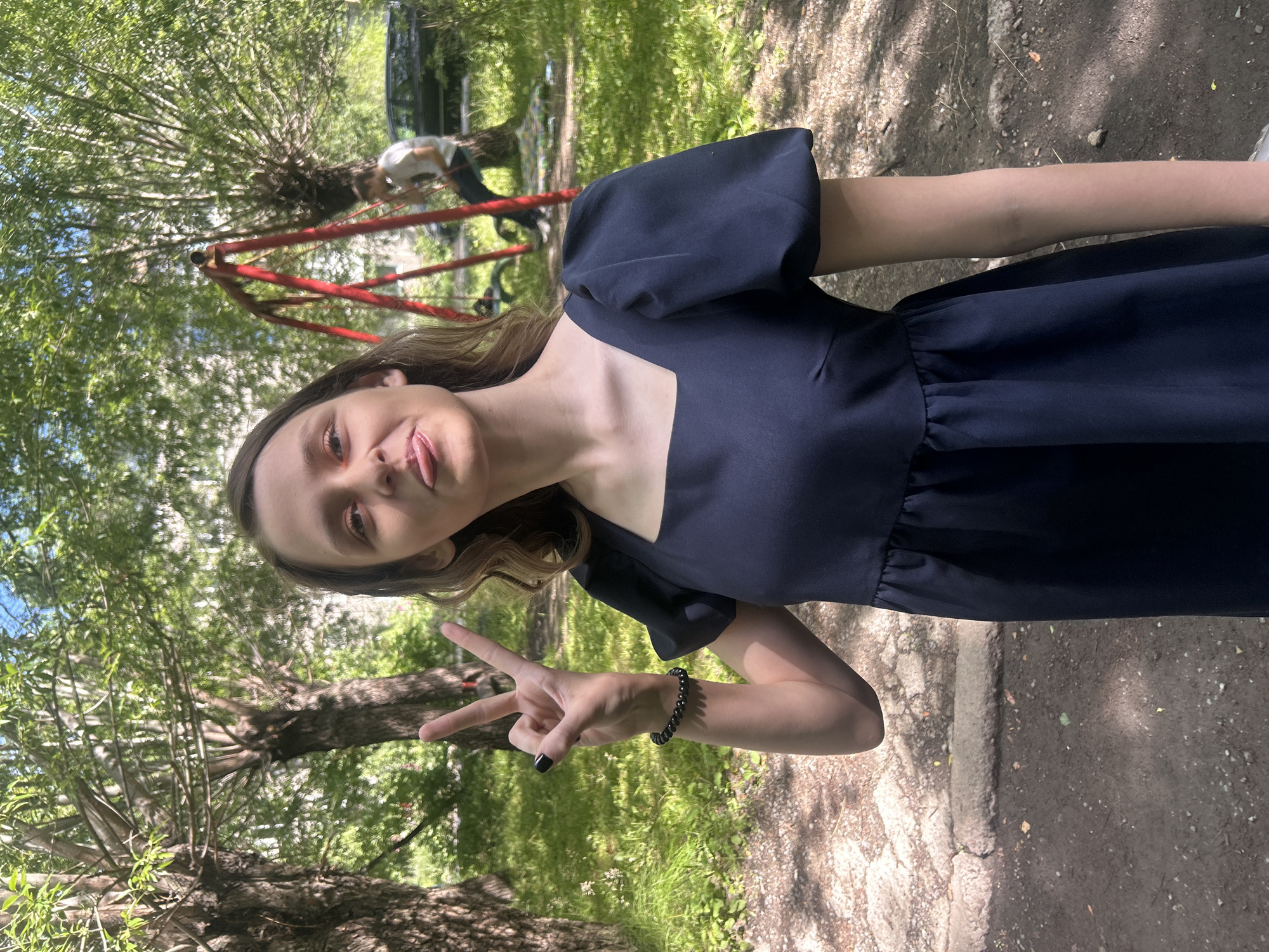 A young woman with light brown hair stands in an outdoor setting, possibly a park or courtyard in Omsk, Russia. She is wearing a dark blue dress with puff sleeves and a square neckline. Her left hand is raised, forming a peace sign, with dark nail polish. She has a bracelet of black beads on her left wrist. Her facial expression is playful, with her tongue sticking out slightly. Behind her, a red swing set is visible, and a man is sitting on a swing in the background. Trees with green foliage surround the area, and the ground appears to be a mix of dirt and asphalt. The lighting suggests it is daytime.