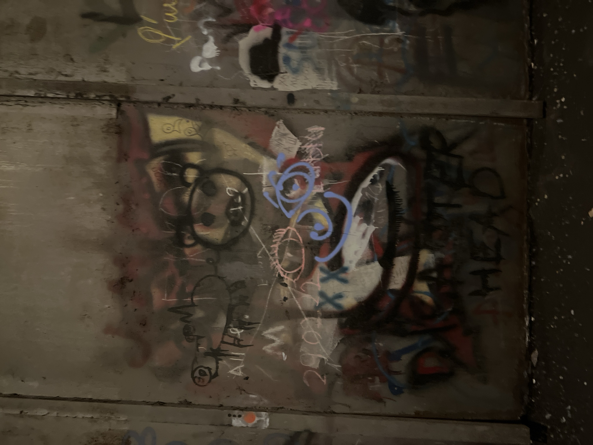 The image shows a close-up of a concrete or metal wall covered in graffiti. The graffiti includes various tags, symbols, and abstract shapes in different colors such as black, blue, pink, white, and red. The surface appears weathered and layered with overlapping artwork. The right edge of the image shows a darker surface with white speckles, possibly the wall or edge of a structure.
There is no verifiable information about the exact location in Yerevan, Armenia, or any specific people, actions, or interactions. The subject is solely the graffiti-covered wall.