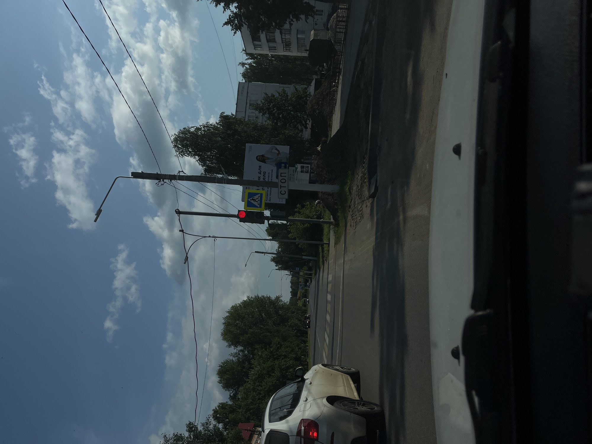 The image captures a view from inside a vehicle, looking forward on a street in Chaikovsky, Russia. The road is paved, with a pedestrian crossing visible. A white SUV is in the left lane, and other vehicles are further down the road. Traffic lights are present, with a red light illuminated. A stop sign is also visible. Buildings line the right side of the street, with trees and a cloudy blue sky overhead. There are several power lines and streetlights along the road. Construction debris is piled up on the right side of the street.