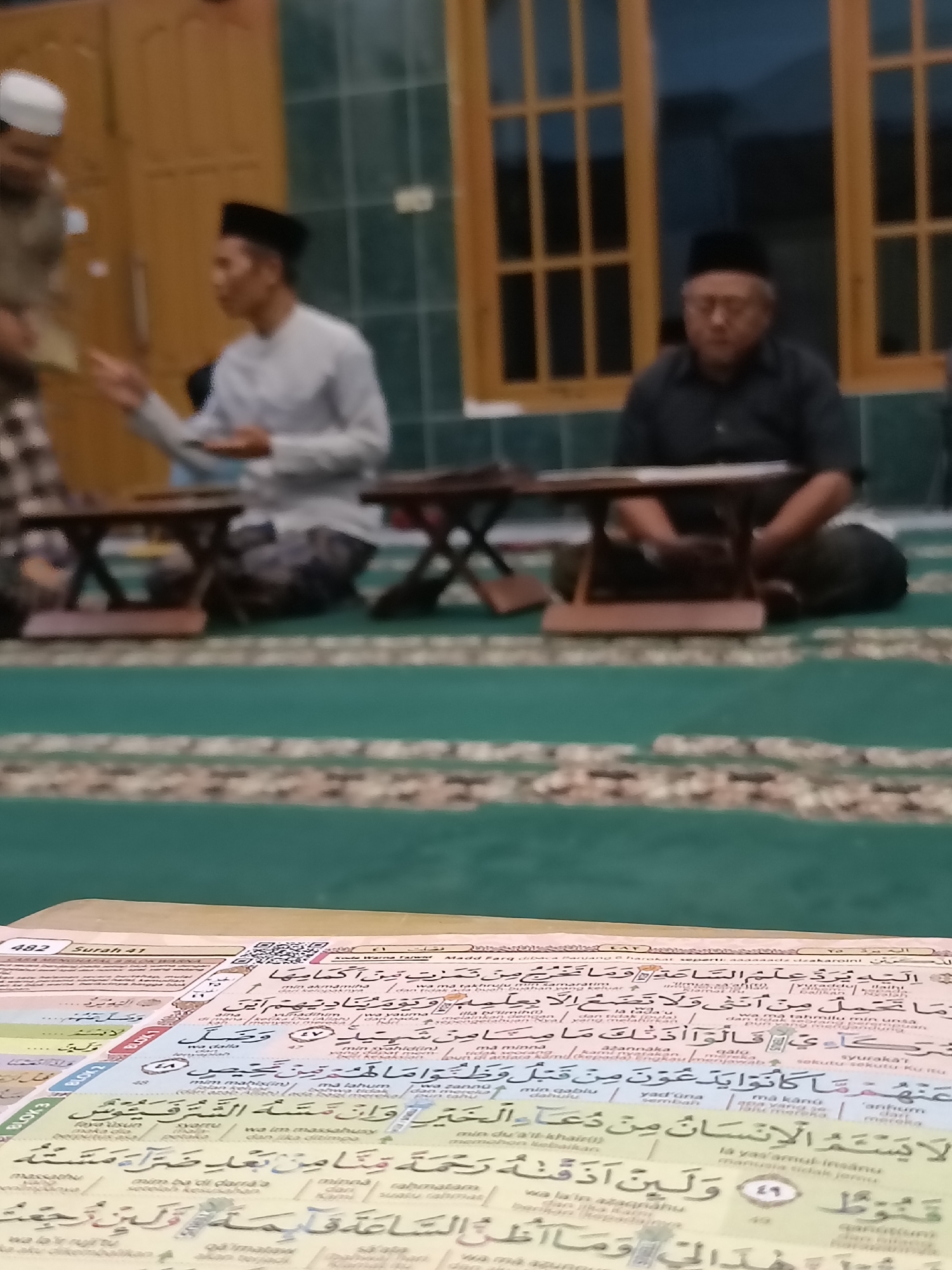 The image shows a group of Muslim men in traditional attire sitting on a green carpet in what appears to be a prayer room or a religious gathering space. Two men are visible in the foreground, seated on the floor with small wooden tables in front of them. One man is wearing a white shirt and a black cap, while the other is wearing a dark shirt and a black cap. A third man, wearing a white cap, is partially visible in the background. The men appear to be engaged in a religious activity, possibly reading or studying religious texts, as a book, likely the Quran, is open in the foreground. The environment is dimly lit, with wooden doors and windows visible in the background. The carpet has a patterned border. The image is captured in Kediri City, Indonesia, indicated by the presence of the Quran and the traditional Muslim attire of the individuals, consistent with the cultural context of the location.