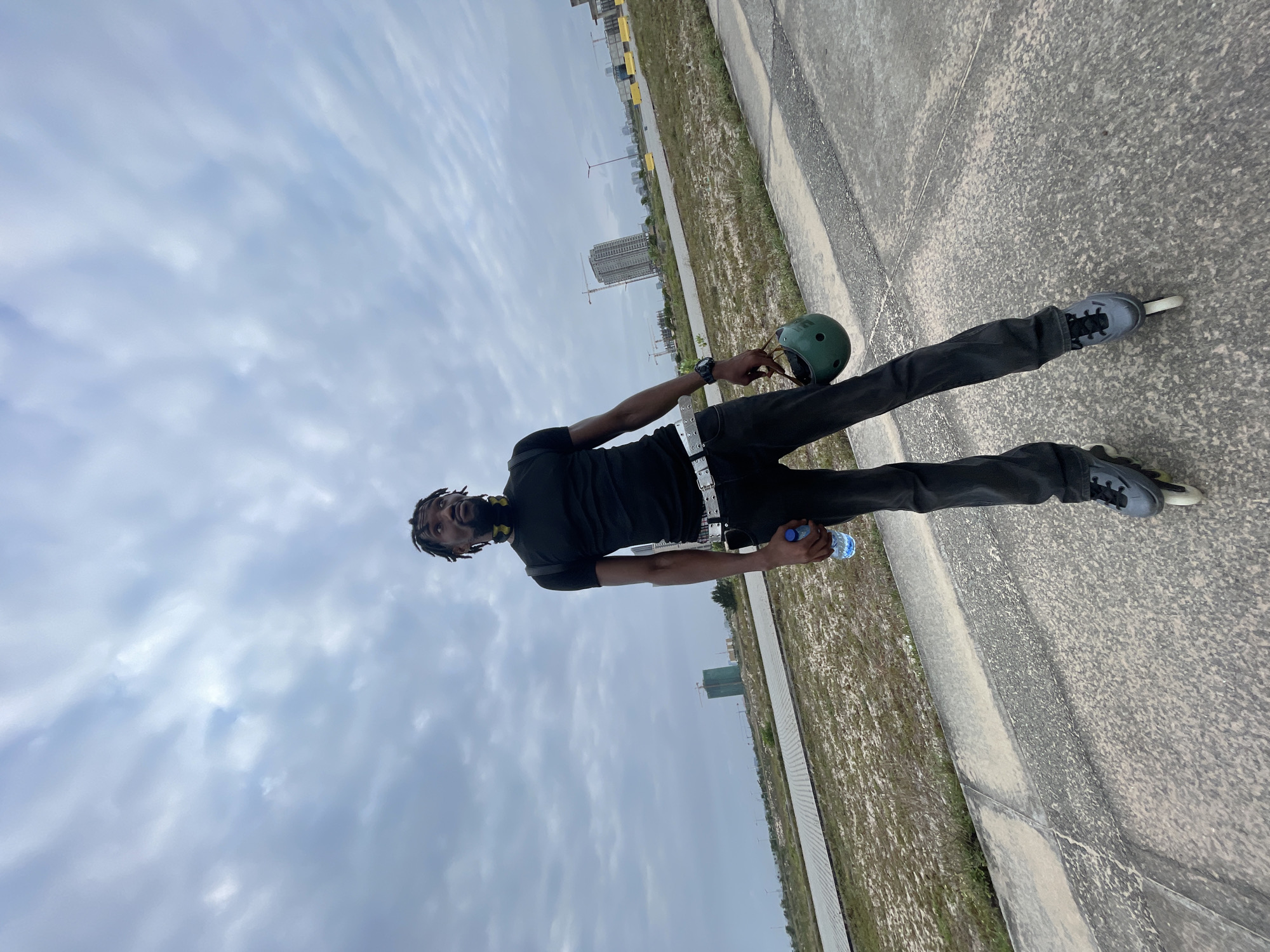 A full-body, low-angle shot captures a Black man in rollerblades standing on a paved path in Beecroft Point, Nigeria. He wears a black t-shirt, black pants, and a white belt. In his right hand, he holds a green helmet, and in his left, a bottle of water. The background features a vast, open area with scattered vegetation, construction sites with cranes, and distant buildings under a cloudy sky. The man is looking upwards and to his right, his expression thoughtful.