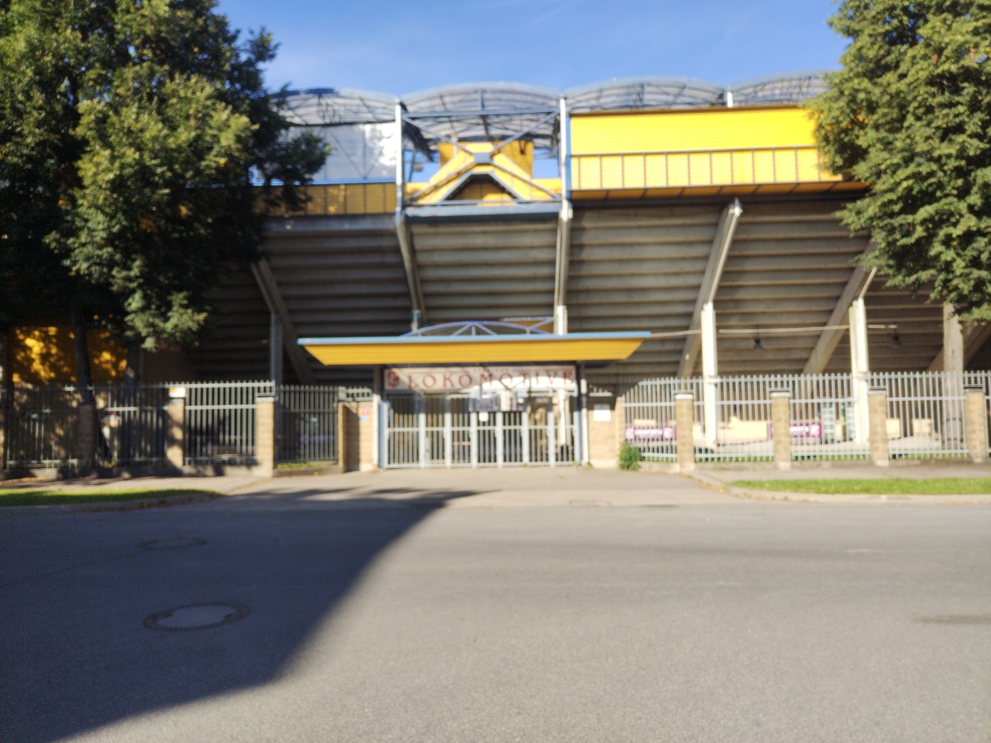 The image shows the entrance to the Daugavpils Olympic Center "Lokomotiv" stadium in Daugavpils, Latvia. The stadium features prominent yellow and grey structures, with tiered seating visible behind a metal fence. A sign above the main entrance reads "LOKOMOTIVE". Trees line the perimeter of the stadium, and a street with manhole covers is in the foreground. The sky is blue and clear. There are no people or vehicles visible in the image. The overall scene depicts an outdoor sports facility under bright daylight.