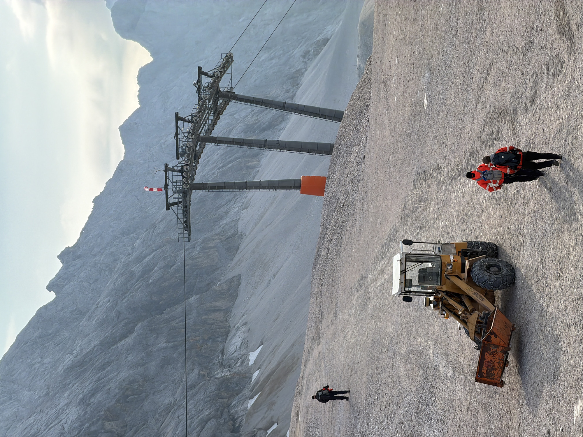 This image depicts a mountainous, rocky terrain, likely in the Bavarian Alps near Garmisch-Partenkirchen, Germany. In the foreground, a yellow front-end loader with an orange bucket is parked on a gravel path. Two individuals wearing red jackets and backpacks are walking away from the camera, also on the gravel path. Further in the background, another person wearing a dark jacket and a backpack is walking towards the left side of the frame. A ski lift structure with multiple pylons and cables is visible in the mid-ground, with a large orange padded protector on one of the pylons. The background features a vast, steep, and rocky mountain face with patches of snow, under an overcast sky.