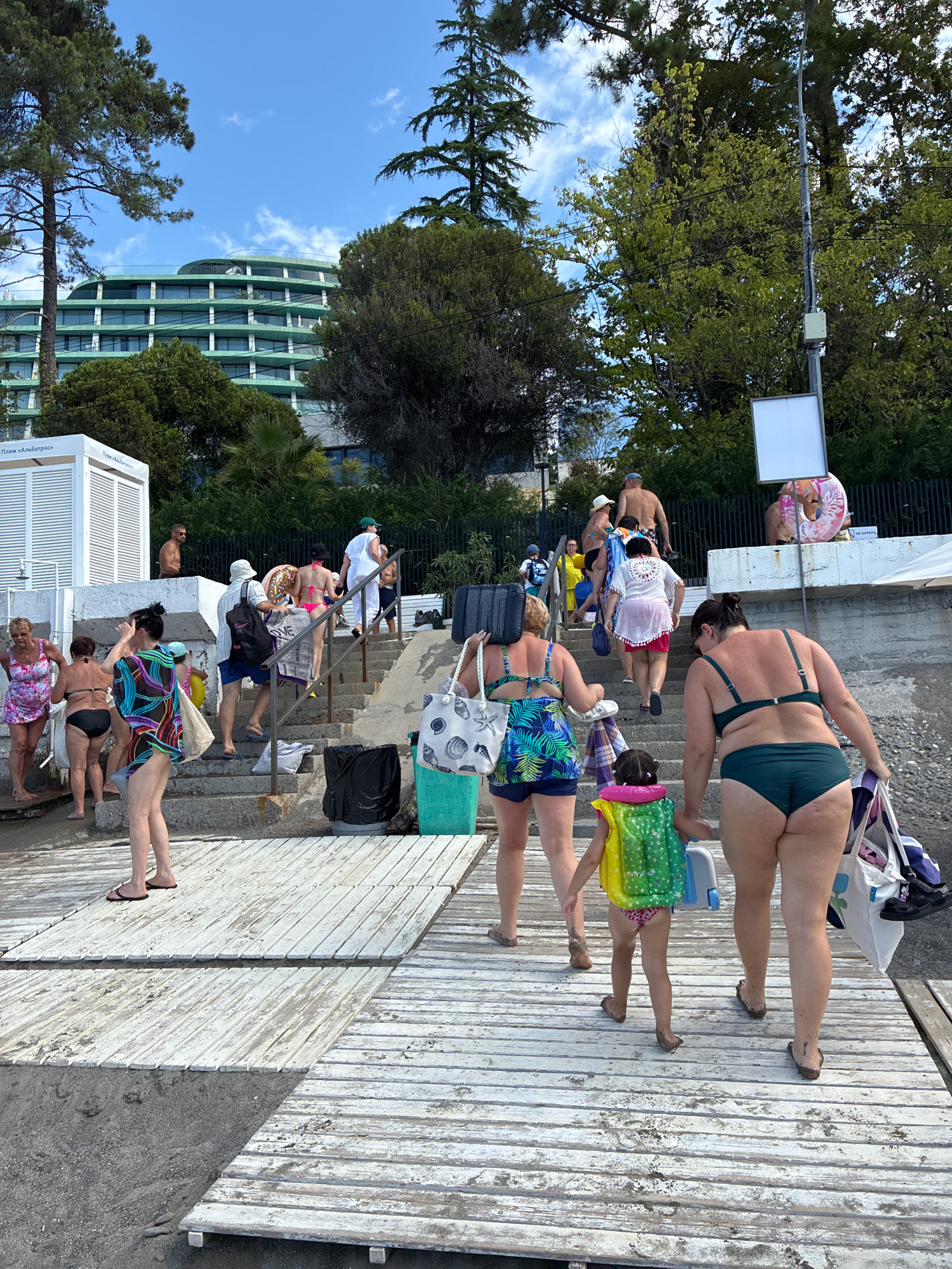 The image depicts a group of people ascending concrete stairs on a beach, likely in the city of Sochi, Russia, given the context of a resort hotel visible in the background. The hotel is a large, modern structure with a distinctive green, tiered facade, suggesting it is a prominent beachfront establishment. A wooden boardwalk is visible in the foreground, with some people walking on it before reaching the stairs.
Several individuals are dressed in swimwear, carrying various beach accessories such as bags, a suitcase, and a cooler. One woman wears a colorful patterned swimsuit and a matching cover-up. Another woman is in a dark green bikini. A young child in a life vest is also present, holding hands with an adult.
The environment appears to be a sunny day, with blue skies and scattered clouds. Lush green trees surround the hotel and extend down towards the beach area. The stairs are flanked by a low wall on one side and dense foliage on the other. Two trash cans, one black and one green, are positioned near the base of the stairs. A circular inflatable ring is visible on the wall further up the stairs, alongside a white sign.
The scene conveys a sense of everyday life at a beach resort, with people returning from or heading to the sea. The overall impression is one of leisure and recreation.