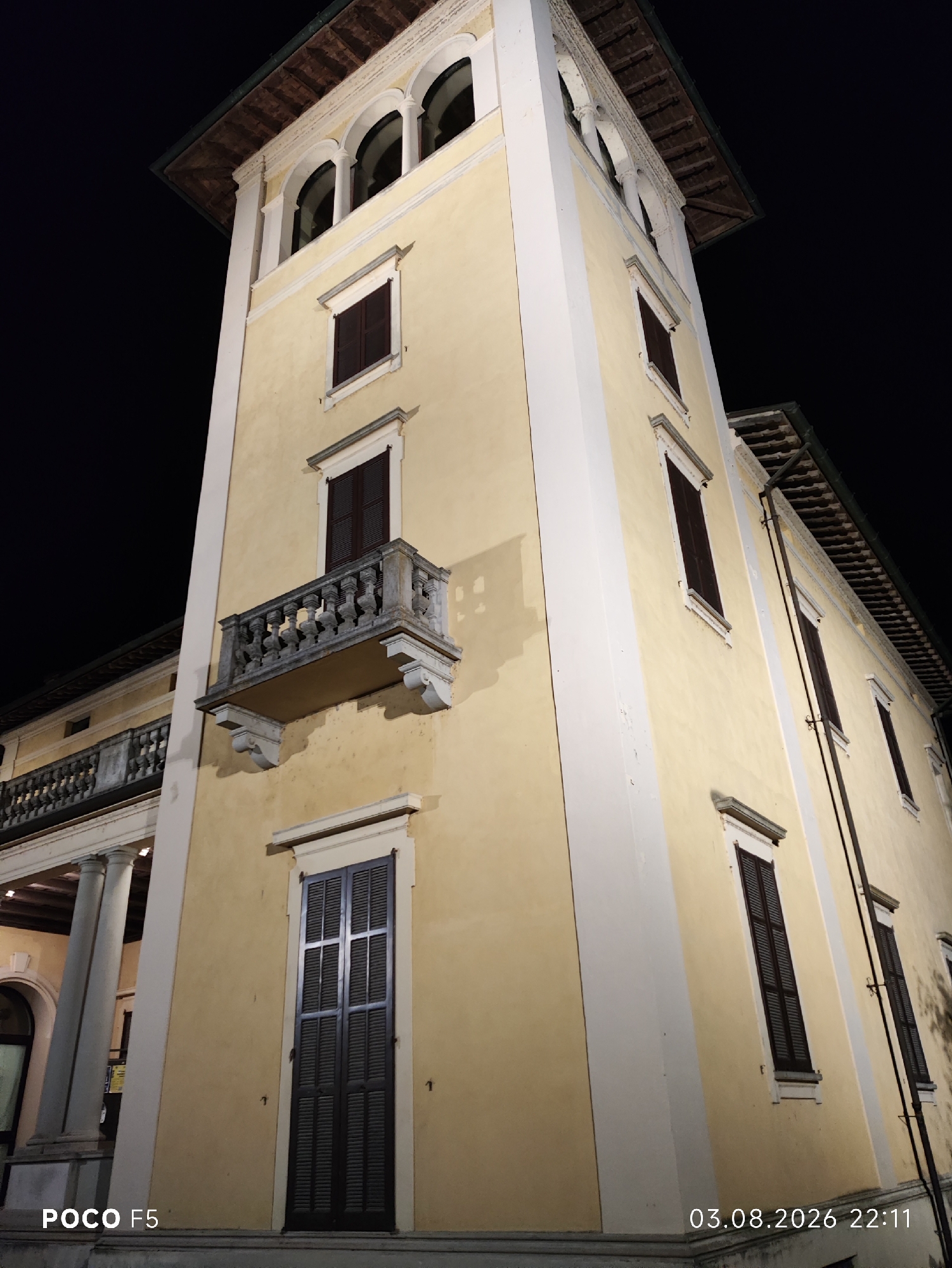 A low-angle, nighttime photograph captures the side of a pale yellow, multi-story building with a dark tiled roof, illuminated by artificial light. The building features several arched windows at the top, resembling a loggia, with a dark tiled roof extending outwards. Below this, on the main facade, are several shuttered windows with dark brown shutters. One prominent feature is a stone balcony with ornate balusters, projecting from the second floor. The building has white trim around the windows and along the corners. In the foreground, the bottom section of the building has a large, tall shuttered window. The lower left corner of the image displays the text "POCO F5," and the lower right corner shows the date and time "03.08.2026 22:11." The surrounding environment is dark, indicating nighttime. The building appears to be a historical structure, possibly a villa or public building, situated in the city of Collecchio, Italy.
