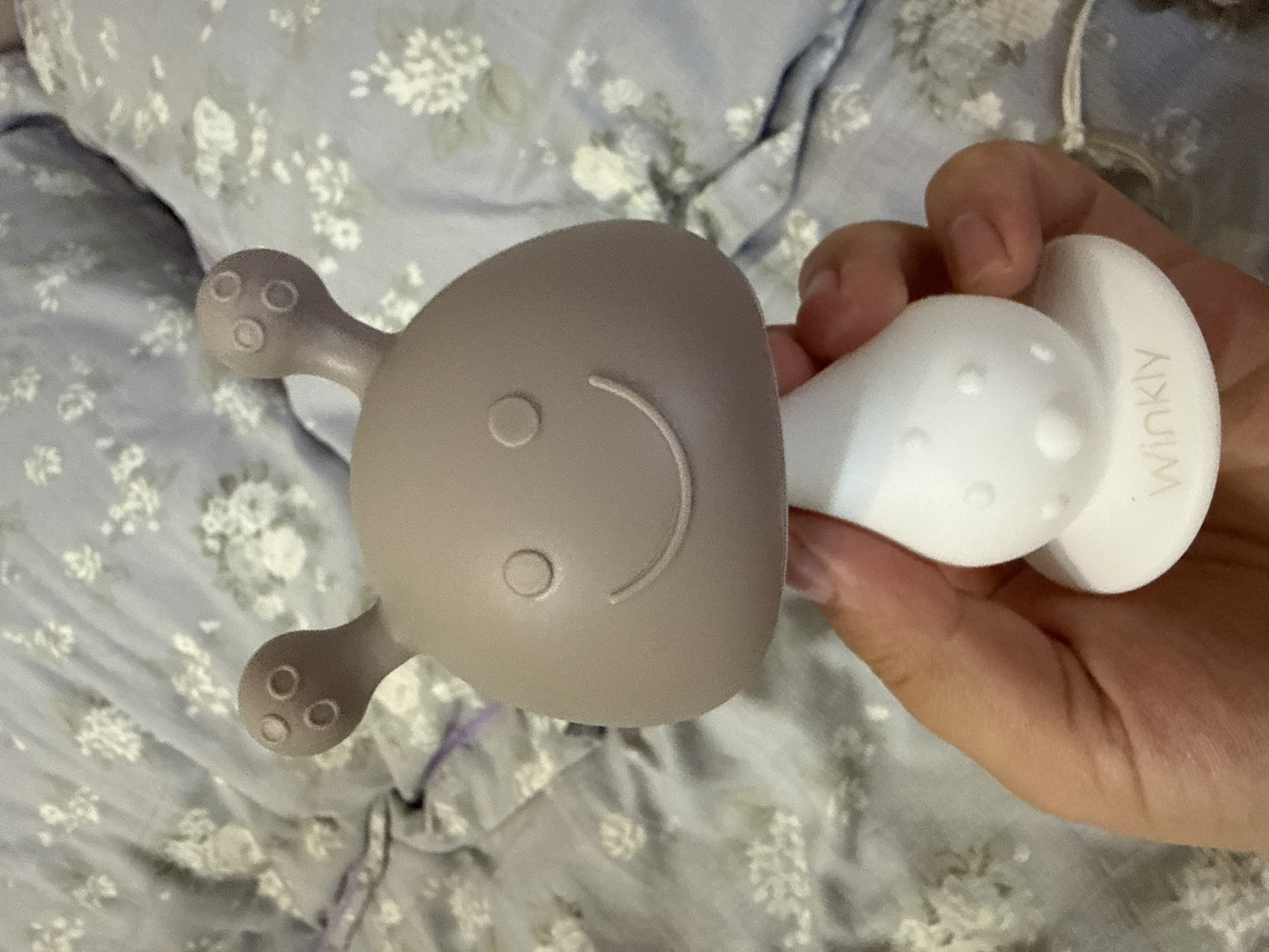A hand holds a two-toned baby teether in front of a floral-patterned blanket. The teether has a grey, mushroom-shaped top with two antennae, each with three small circles, and a smiley face made of a circle and a curved line. The bottom half of the teether is white and bulbous, with raised bumps, and a circular base. The word "Winkly" is embossed on the base. The blanket is light blue with white floral designs.
