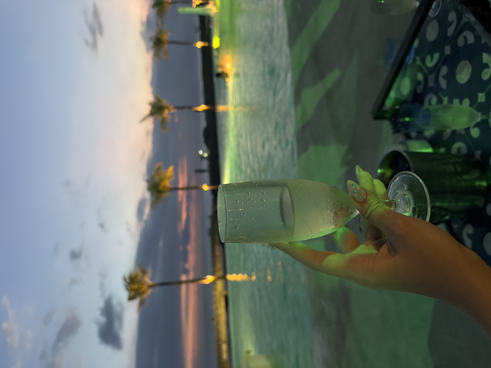 This image captures a serene twilight scene in North Kuta, Indonesia, likely at a resort or hotel. In the foreground, a hand, adorned with light-colored fingernails featuring small embellishments, elegantly holds a champagne flute. The glass is filled with a clear liquid and has condensation on its exterior, suggesting a cold beverage.
Beyond the glass, the backdrop features a beautiful dusk sky with soft hues of light blue, pink, and orange near the horizon, accented by scattered clouds. Several palm trees are silhouetted against the colorful sky, their shapes reflected in the calm waters below. A large body of water, possibly a swimming pool or a decorative lagoon, dominates the midground. This water is strikingly illuminated with a vibrant green light, creating a glowing, atmospheric effect.
To the right of the frame, a dark table or surface with a distinctive patterned tablecloth (black with green or white circular motifs) is visible, along with parts of another glass. The green lighting from the water casts a luminous glow over the table and the hand holding the drink. The scene evokes a sense of relaxation and luxury, typical of an evening at a tropical destination. There is no visible text in the image.