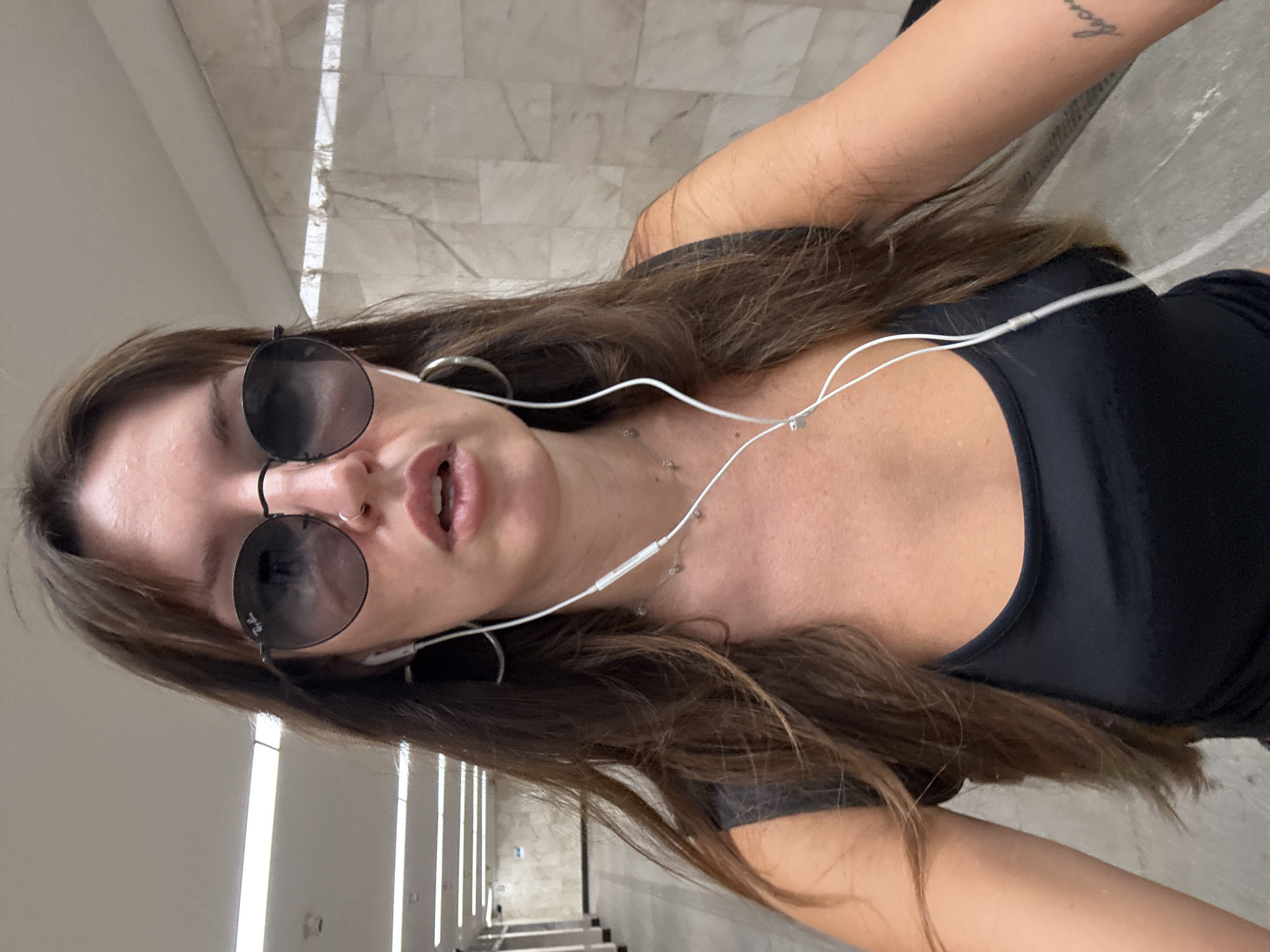 A woman with long, dark brown hair, wearing sunglasses and earbuds, stands in a subway station. She is wearing a black tank top and a silver necklace. A tattoo is visible on her right forearm. The station features marble-tiled walls and fluorescent lighting. The image was taken in a Moscow Metro station.