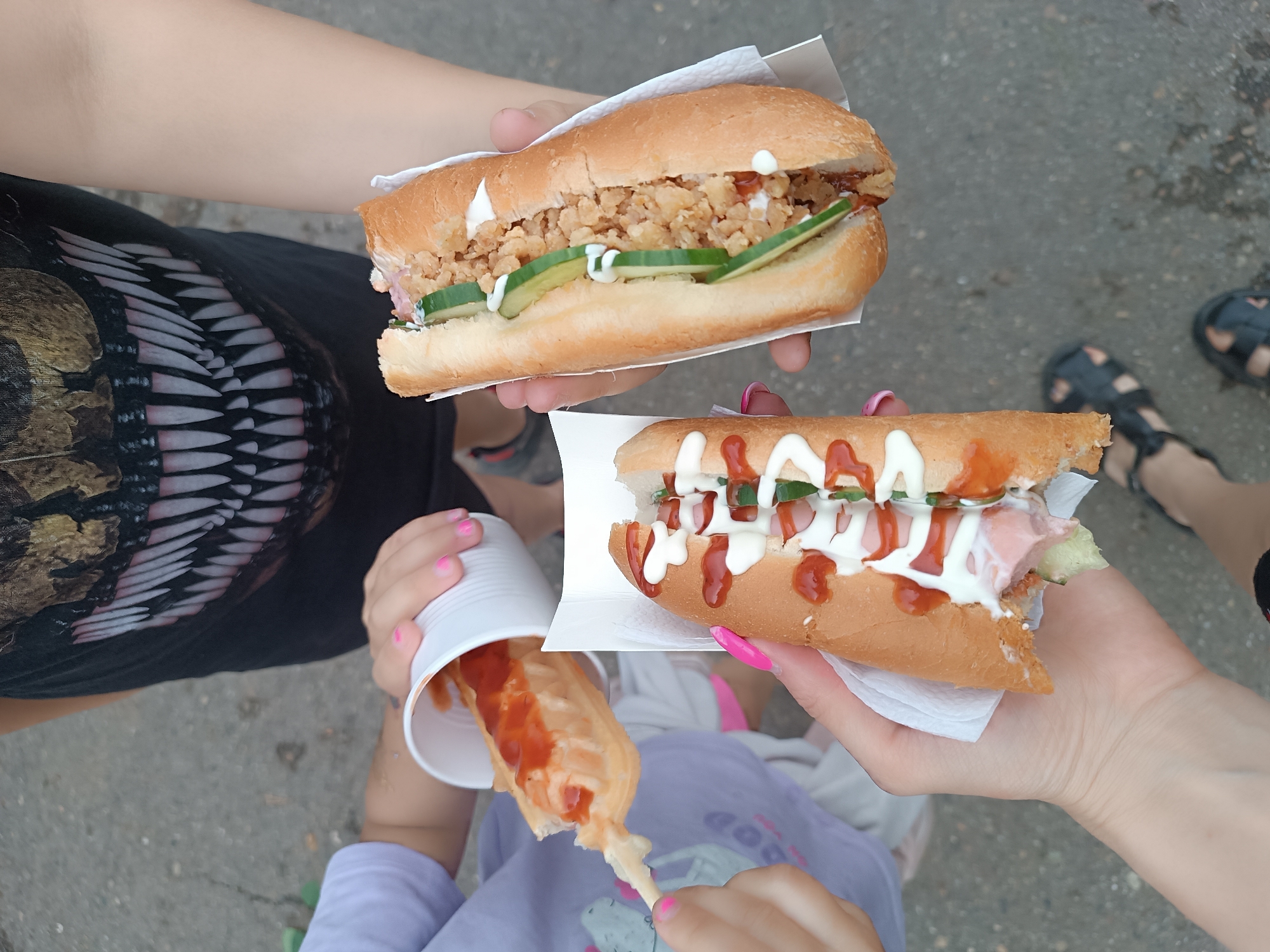 Three people are pictured from a high angle, holding food. One person is holding a long sandwich filled with breaded and fried material and cucumber slices, dressed with mayonnaise. A second person holds a similar sandwich filled with ham, cucumber slices, mayonnaise, and ketchup. A third person holds a corn dog on a stick in a white cup, with ketchup applied. The setting is outdoors on a paved surface. The location is most likely Chaikovsky, Russia.