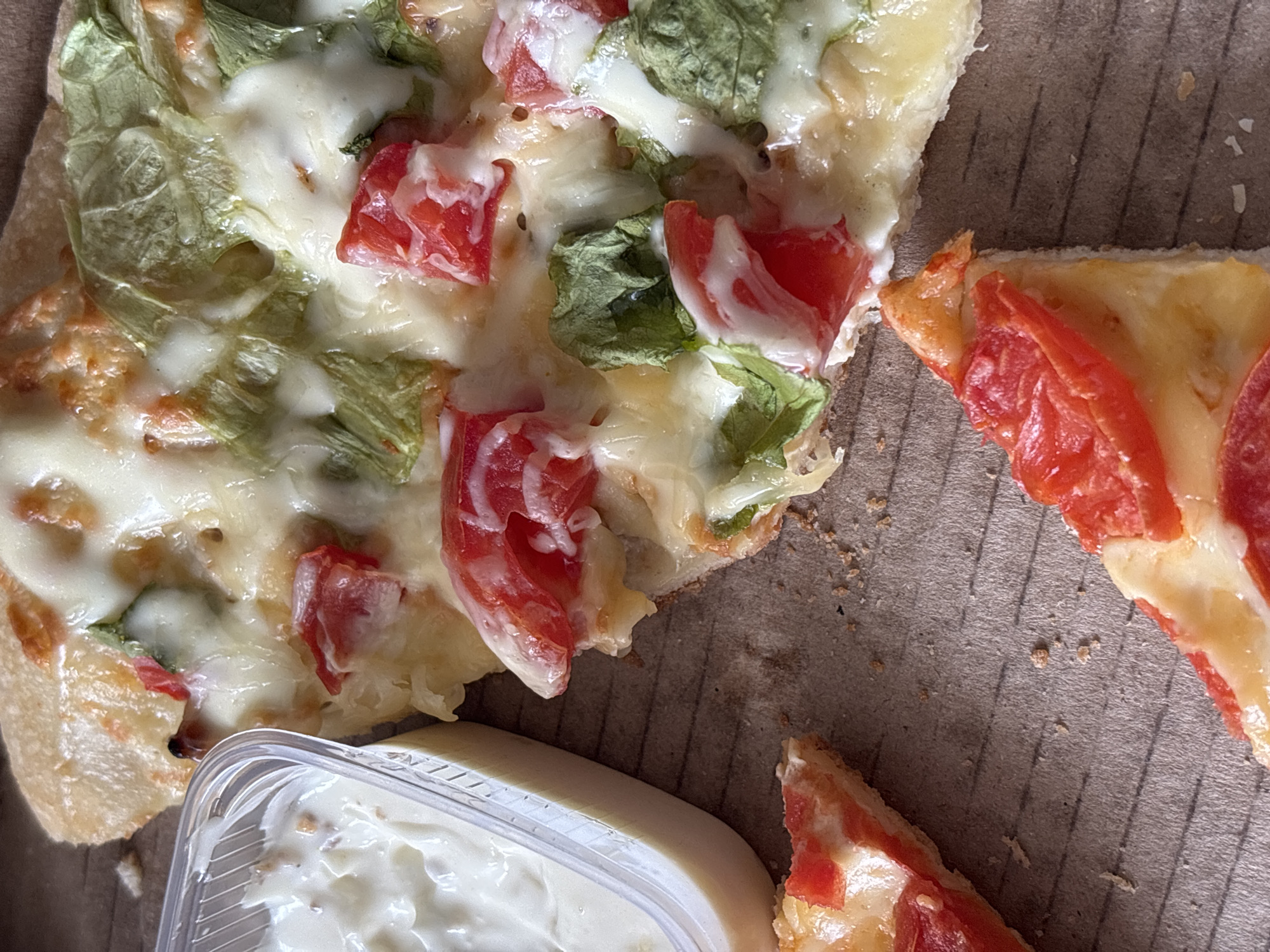 The image shows leftover pizza in a cardboard box, possibly in Perm, Russia. A container of white sauce sits near a large piece of pizza topped with cheese, tomatoes, and green lettuce. Nearby, another smaller piece of pizza has tomato slices on top. The interior of the cardboard box is a plain brown color. There are crumbs scattered in the box.