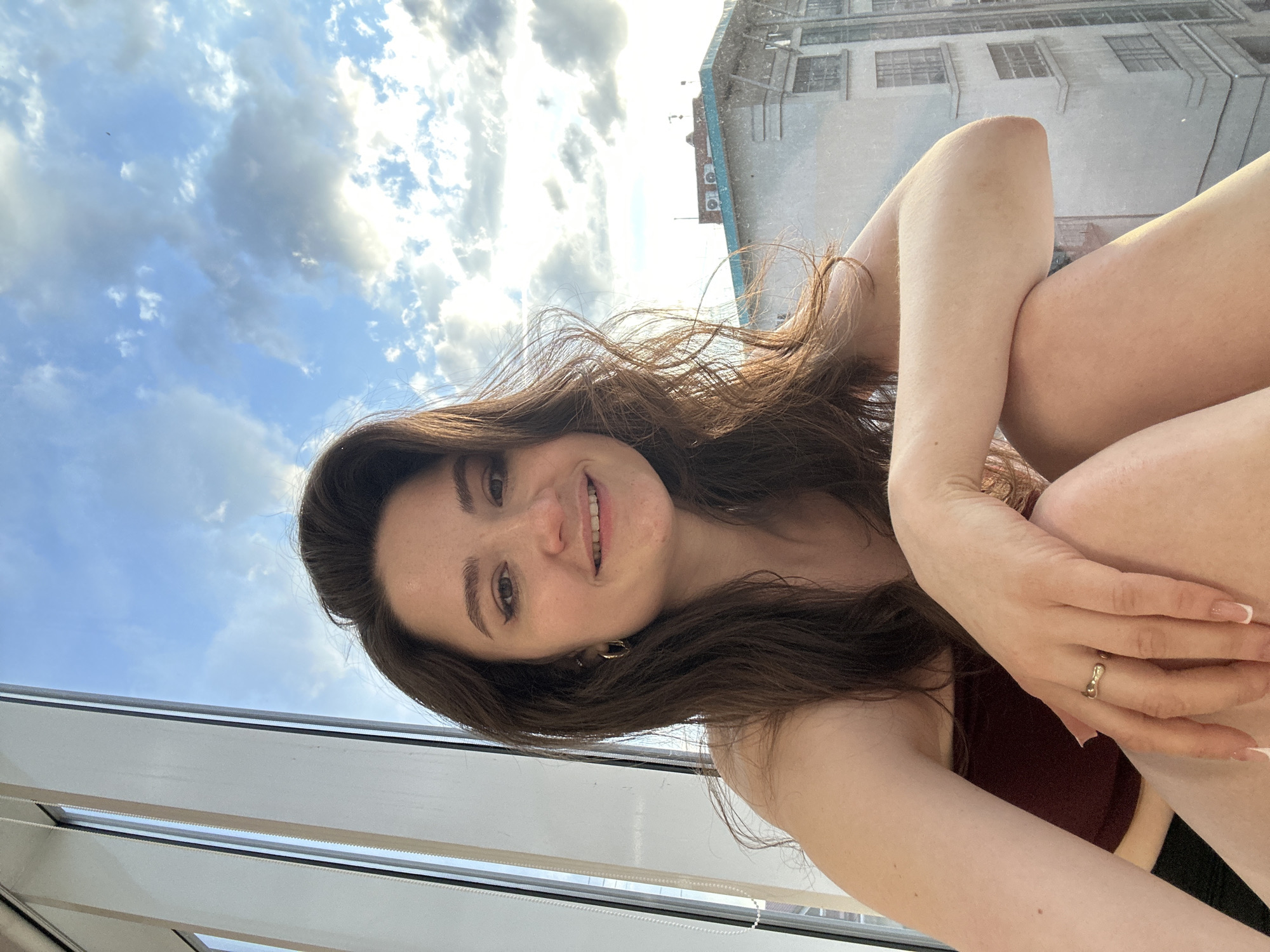 A portrait-oriented photograph captures a young woman smiling brightly at the camera, taken from a low angle. She has long, flowing dark brown hair and appears to be sitting or crouching, with her arms wrapped around her knees. Her head is slightly tilted, and she gazes directly at the viewer with a pleasant, happy expression. She is wearing a dark top, possibly maroon or dark red. A golden ring adorns her left ring finger, and her fingernails are styled with a white-tipped French manicure.
The background features a clear blue sky with scattered white clouds, some brightly illuminated by the sun, suggesting daytime, possibly late afternoon or early evening. To the right of the woman, an urban landscape is visible, including an older building with multiple windows and a distinct light blue architectural feature with what appears to be air conditioning units. A white window frame or railing is partially visible at the bottom of the image, indicating the woman is positioned near a window or balcony overlooking the city. The setting is Ufa, Russia. No text is visible in the image.