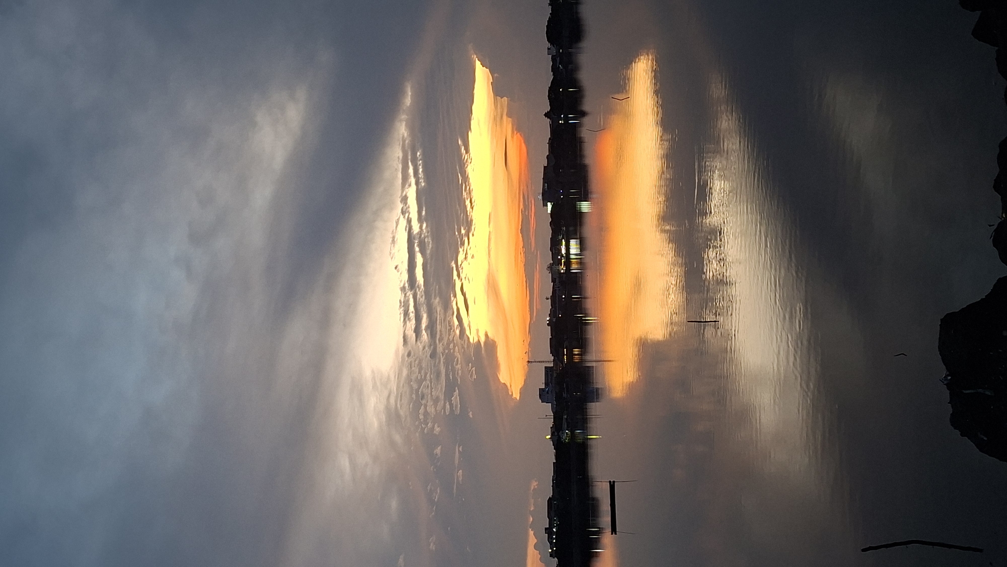 The image captures a serene sunset over the waters of Lhokseumawe, Indonesia. The sky is a dramatic display of colors, with dark, heavy clouds in the upper portion transitioning to vibrant oranges and yellows near the horizon, where the sun is setting. A distinct band of bright orange light illuminates the underside of a large, wispy cloud formation, creating a striking visual contrast against the darker sky.
Below the sky, a silhouetted skyline of buildings and structures stretches across the horizon. These buildings appear to be of moderate height, with some taller antenna-like structures visible. Lights from the buildings twinkle, suggesting a populated area.
The water in the foreground is calm and reflects the dramatic colors of the sky and the illuminated buildings. The reflection is a mirror image, though slightly distorted by gentle ripples on the water's surface. A small, dark, rectangular object, possibly a boat or a buoy, is visible in the left middle ground of the water. Several thin, dark vertical sticks or posts emerge from the water at various points, adding to the textured detail of the scene. The lower edge of the image shows a dark, rocky foreground, partially obscuring the water's edge. The overall impression is one of tranquility and the beautiful spectacle of nature at dusk.