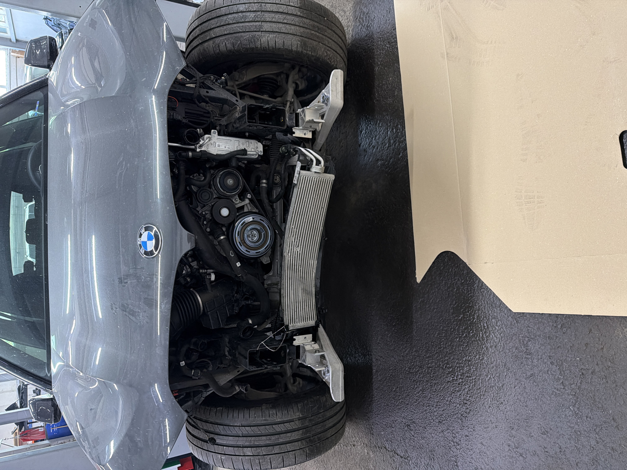 The image captures the front section of a gray BMW car undergoing repair or maintenance in an automotive workshop. The car's front bumper and grille have been removed, exposing the intricate components of the engine bay. Visible elements include the engine block, serpentine belt, various pulleys, and a prominent radiator or intercooler assembly positioned at the front.
Two tires are seen in the frame; one is mounted on the front suspension of the vehicle, and another tire lies on the ground or on a stand next to the car. The vehicle appears to be elevated, likely on a hydraulic lift, as part of the repair process. The floor of the workshop is dark and has a textured, slightly bumpy surface. On the right side of the image, a large piece of beige cardboard or paperboard covers part of the background, possibly for protection or as a neutral backdrop.
A clear BMW logo is visible on the gray front fender of the car. No people are present in the image. The lighting suggests it is daytime. The overall setting strongly indicates an active car repair or service environment, likely in Burgas, Bulgaria, given the provided context.