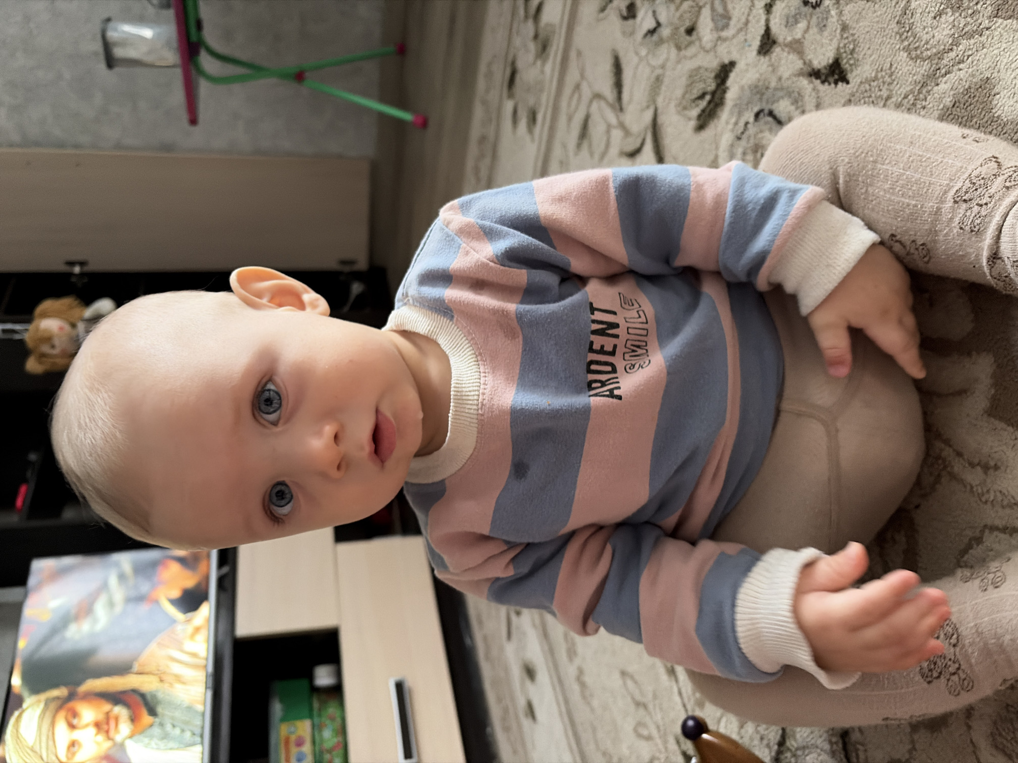A baby with blonde hair and blue eyes is sitting on a patterned rug. The baby is wearing a blue and pink striped sweater with white cuffs and light beige tights. The baby's head is near a dark-colored cabinet with a television on it. A child's stool with pink legs and a green base is visible in the background. The image was taken indoors. The location is in Novopokrovskaya, Russia.