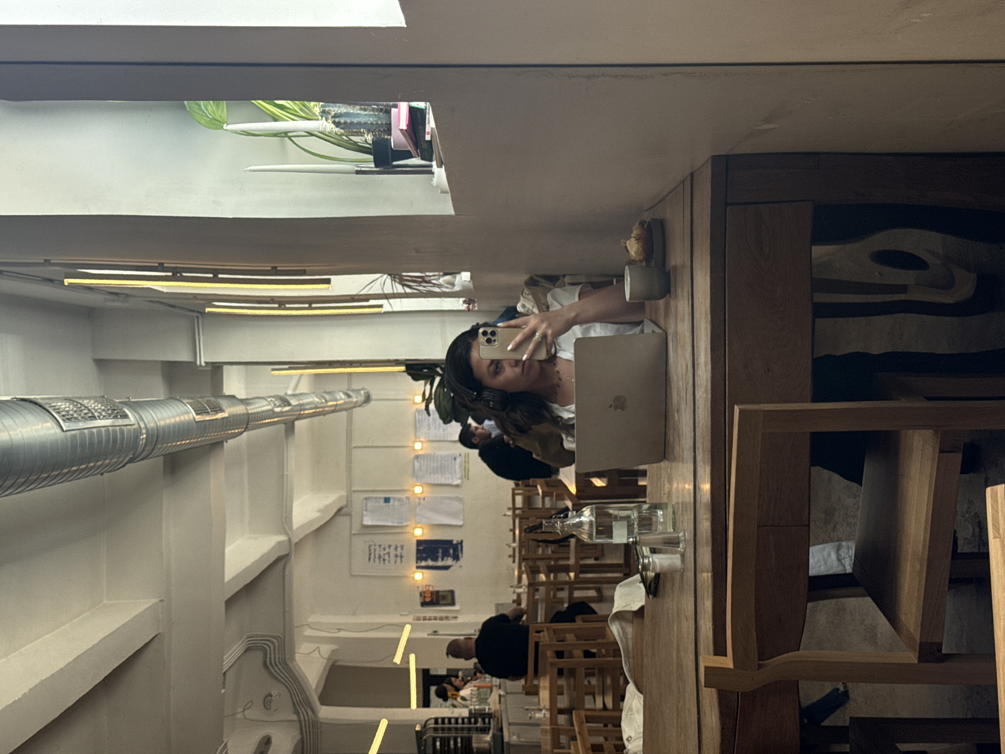 A woman holds a phone while sitting at a wooden table in a restaurant, likely in Sofia, Bulgaria. A laptop is positioned in front of her. The restaurant has rows of tables and chairs leading to the back. Other patrons occupy some of the tables. Light fixtures are present overhead, and decorations line the wall.