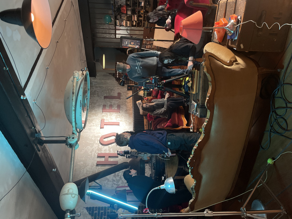 The image depicts an indoor film set operating within a creative space, likely a stylized hotel lobby or lounge, in Paris, France. Six individuals are present, actively engaged in production or awaiting cues. A male in a grey sweatshirt and cap operates a professional video camera mounted on a black tripod, central to the scene. To his left, another male in a dark blue hoodie observes the activity. Further left, a person in a dark hoodie adjusts equipment near a desk, illuminated by a silver floodlight on a stand. In the immediate foreground, a person kneels, arranging items, possibly pastries on a wooden board, atop stacked vintage chests. Two additional individuals are seated on red velvet furniture in the background: one wearing glasses sits on a sofa, and another on an armchair is engrossed in a smartphone. The prominent backdrop features a large 'HOTEL' sign with red letters affixed to a distressed concrete-like wall. Overhead, notable fixtures include a large, vintage industrial multi-arm light and a pendant lamp with an orange interior. A prominent blue LED tube light stands on a tripod to the left, contributing to the scene's cool-toned illumination. An ornate gold-colored chaise lounge rests in the immediate foreground, with coiled black cables visible on the wooden floor.