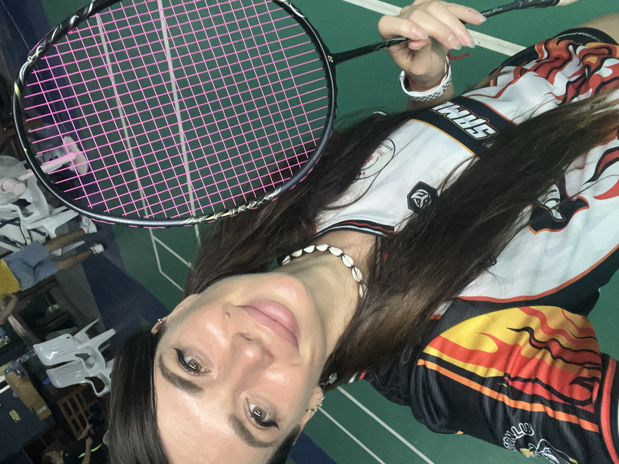 A woman in a white, black, yellow, and red patterned shirt and shell necklace holds a black and silver badminton racket with pink strings. She is indoors on a green court with white lines. The location is unknown.