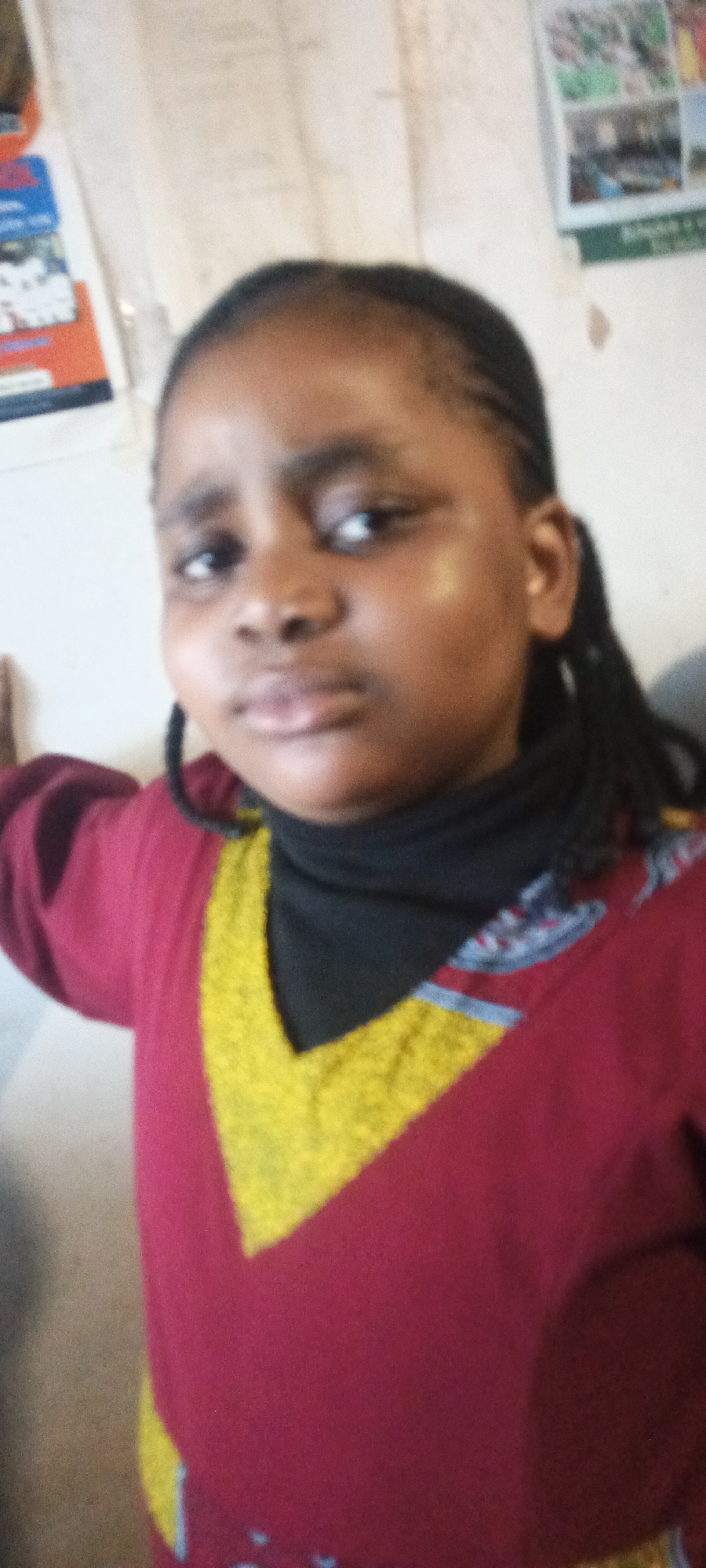 A young Black female is centered in the frame, wearing a black turtleneck under a red top with a yellow V-shaped design. Her hair is styled in braids. The background is a white wall with two posters visible. The location is Unknown, Unknown.
