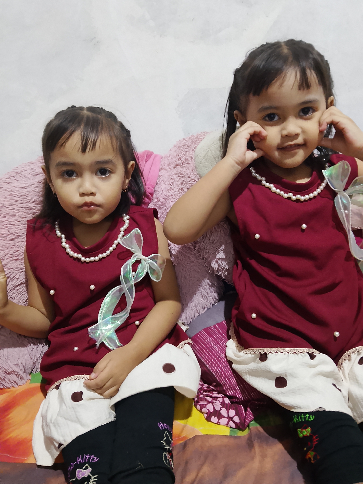 In Tanggungan, Indonesia, two young girls, possibly twins, are pictured sitting on a bed. They are both wearing maroon sleeveless dresses adorned with small white pearls and a decorative ribbon bow on the right side of the younger-looking girl's dress. The dresses have a light-colored hem with dark polka dots. They are also wearing black leggings with "Kitty" and butterfly designs on them. The girl on the left has her hands clasped in front of her, looking at the camera with a neutral expression. The girl on the right is holding her cheeks with her fingers and smiling. They are seated against a white, textured wall, with large pink pillows behind them, suggesting a bedroom setting. The overall impression is a casual, indoor portrait of two children.