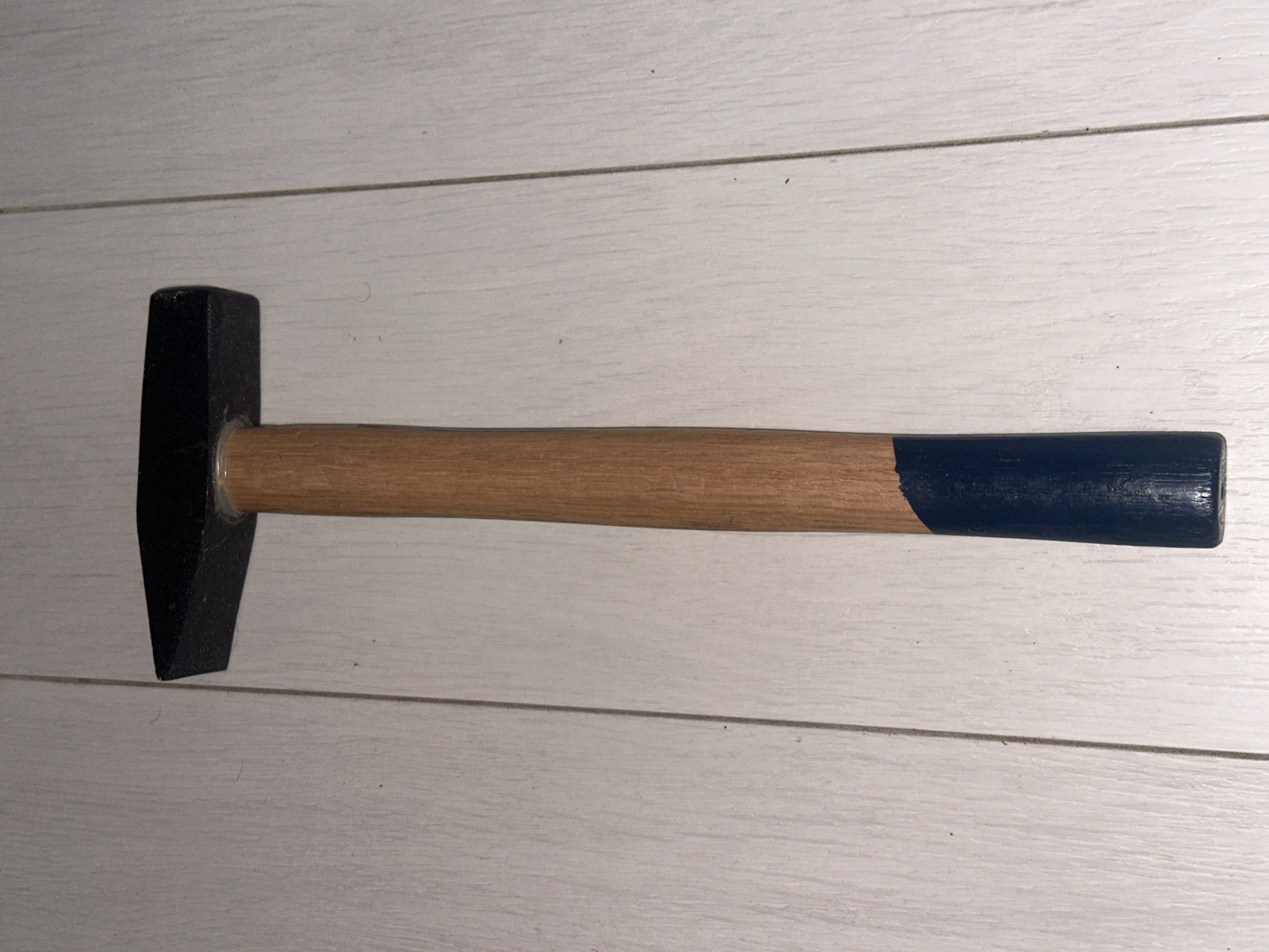 A black hammer with a wooden handle is positioned vertically on a light-colored wooden floor. The handle has a dark blue painted section near the bottom. The scene is a close-up, ground-level shot. There is no indication of the city of Sofia, Bulgaria, or any other specific location.