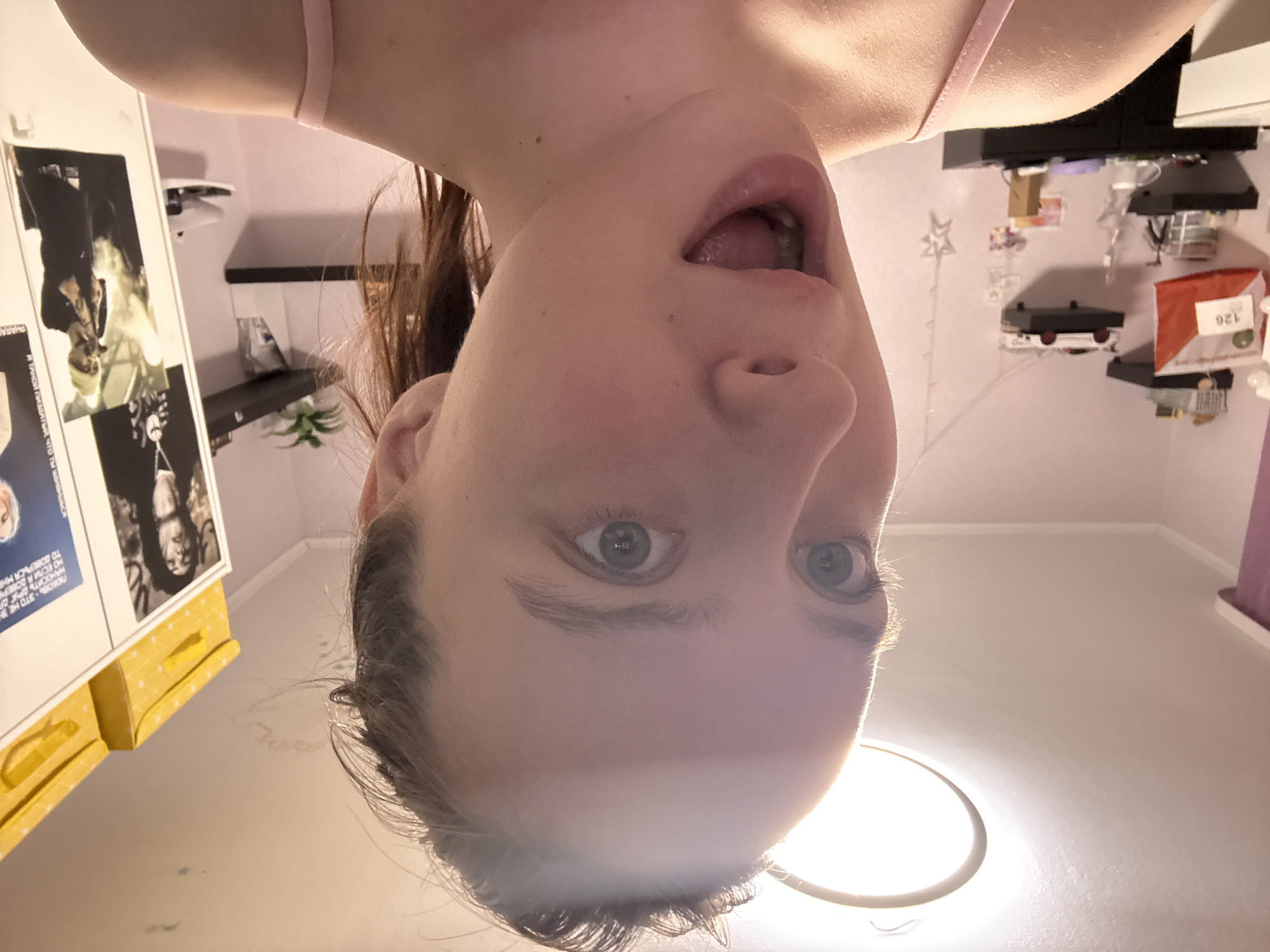 A close-up, upside-down perspective of a person's face. The person, likely a young female, has fair skin, visible grey eyes, and hair pulled back. Their mouth is open in an expression that could be surprise or mild shock. They are wearing what appears to be a light pink tank top.
The background reveals a room interior. A white shelf with various items is visible, including a red package labeled "126." A framed image hangs on the left wall with text in Russian, including the word "ПИЗДАТЫЙ". The ceiling has a circular light fixture. The environment appears to be a personal living space, possibly a bedroom. There is no verifiable information in the image that confirms the exact location as Прикамский, Russia.