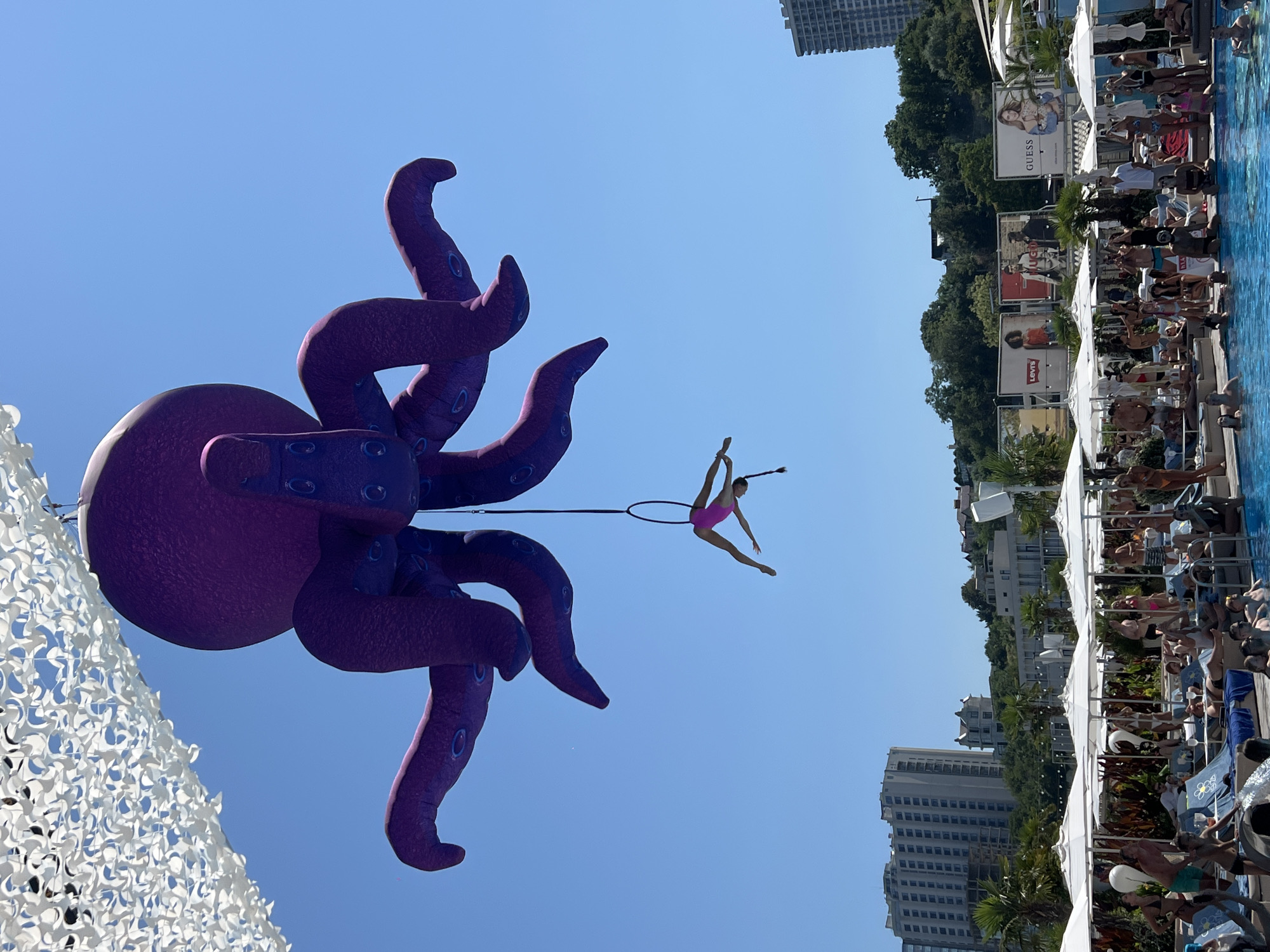 An aerial performer in a pink leotard is suspended from a hoop, performing a backbend against a clear blue sky. The performer is positioned beneath a large, purple, inflatable octopus that hangs from a white canopy structure. The octopus has a textured surface and several visible suckers on its tentacles. Below the performers and octopus, a lively outdoor swimming pool area is visible. Many people are lounging on chairs under white umbrellas, swimming in the pool, or standing around. In the background, buildings, trees, and billboards are present. The scene appears to be a daytime event at a resort or entertainment venue.